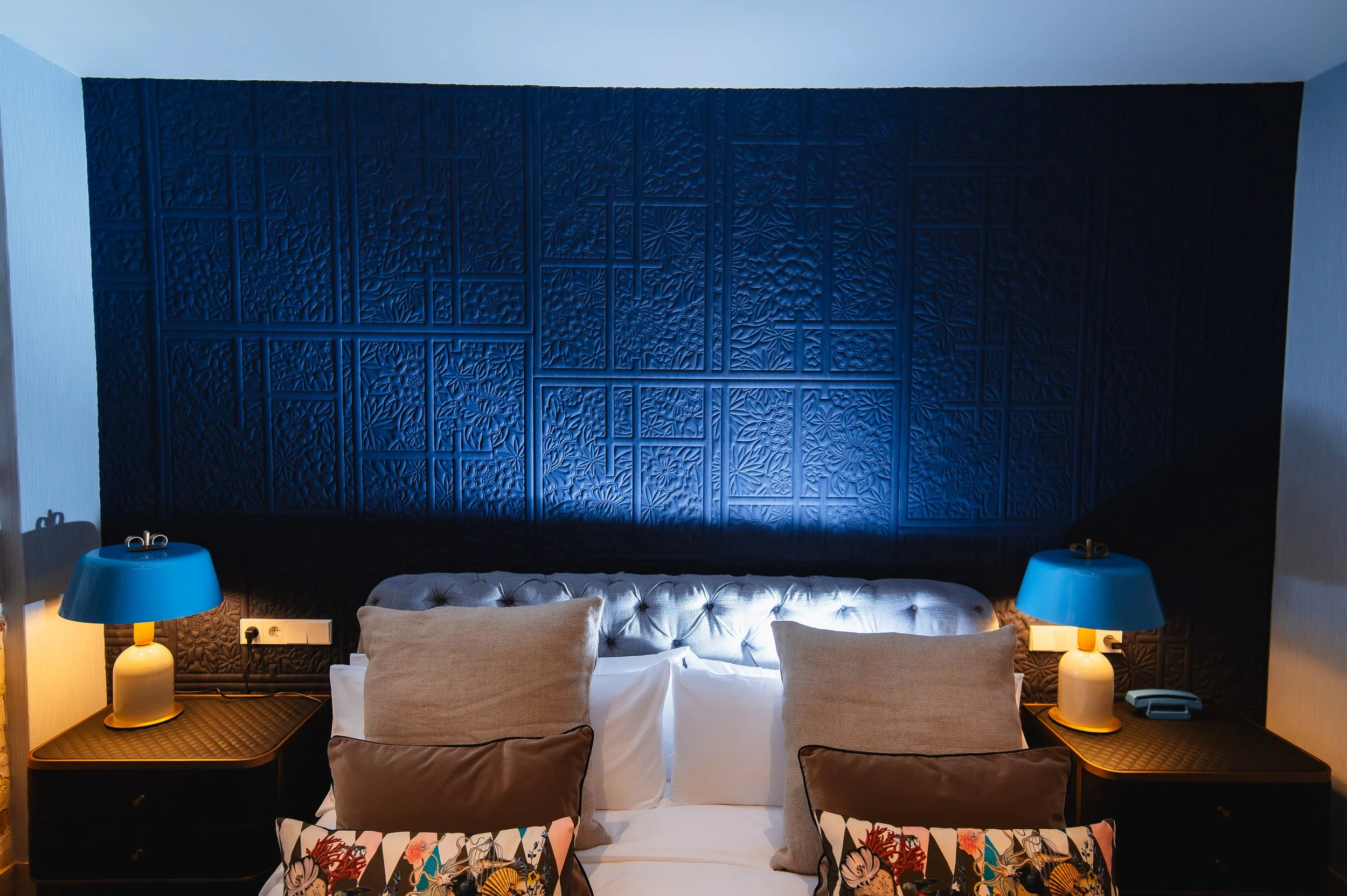 Cama doble en una habitación moderna con cabecera de cuero capitoné, dos mesitas de noche con lámparas azules, y cojines decorativos. La pared de fondo es azul con textura destacada.