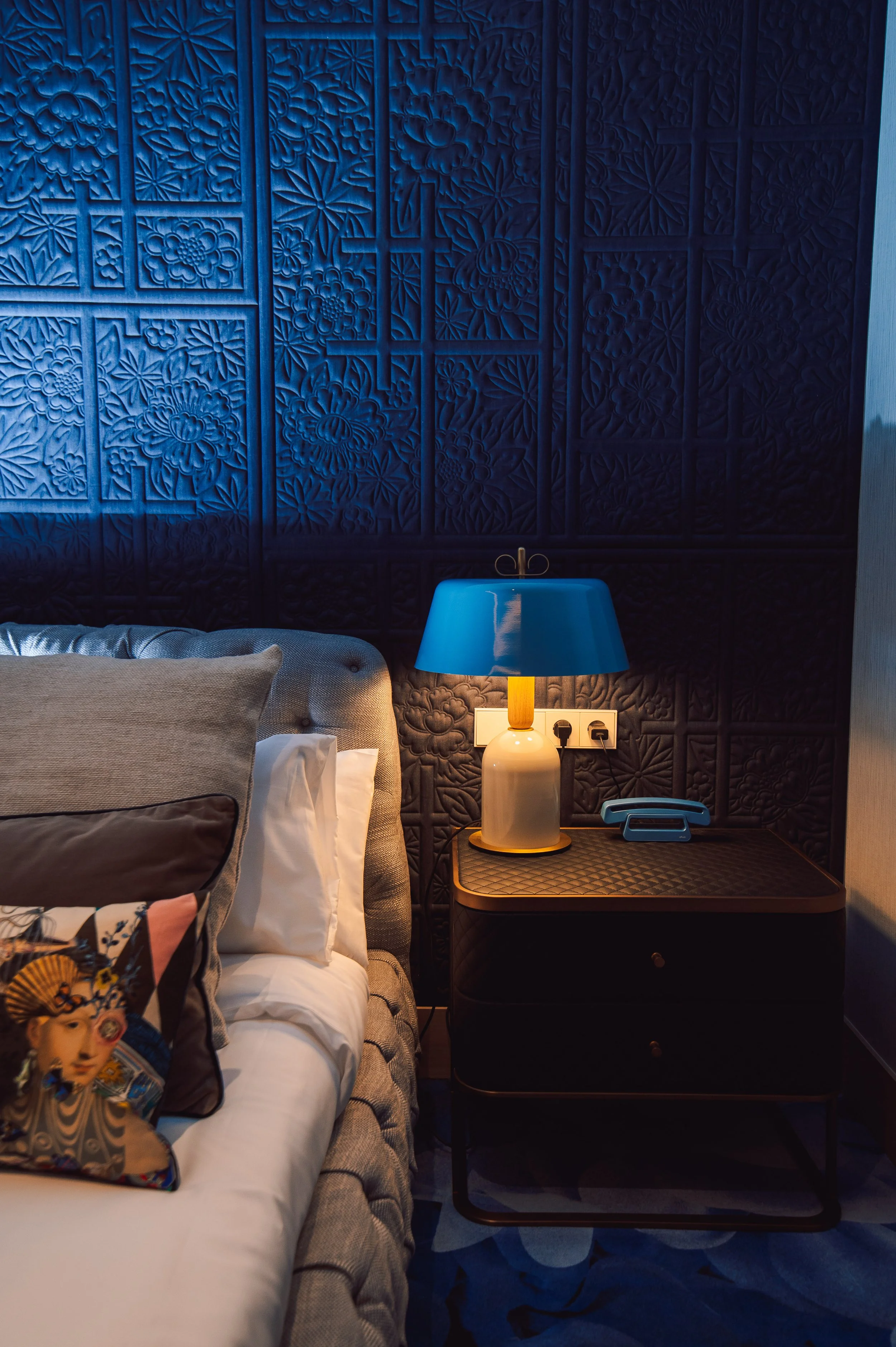 Cabeza de la cama con cojines decorativos, mesa de noche con lámpara azul y teléfono, pared de fondo con textura de paneles con motivos florales en color azul oscuro.