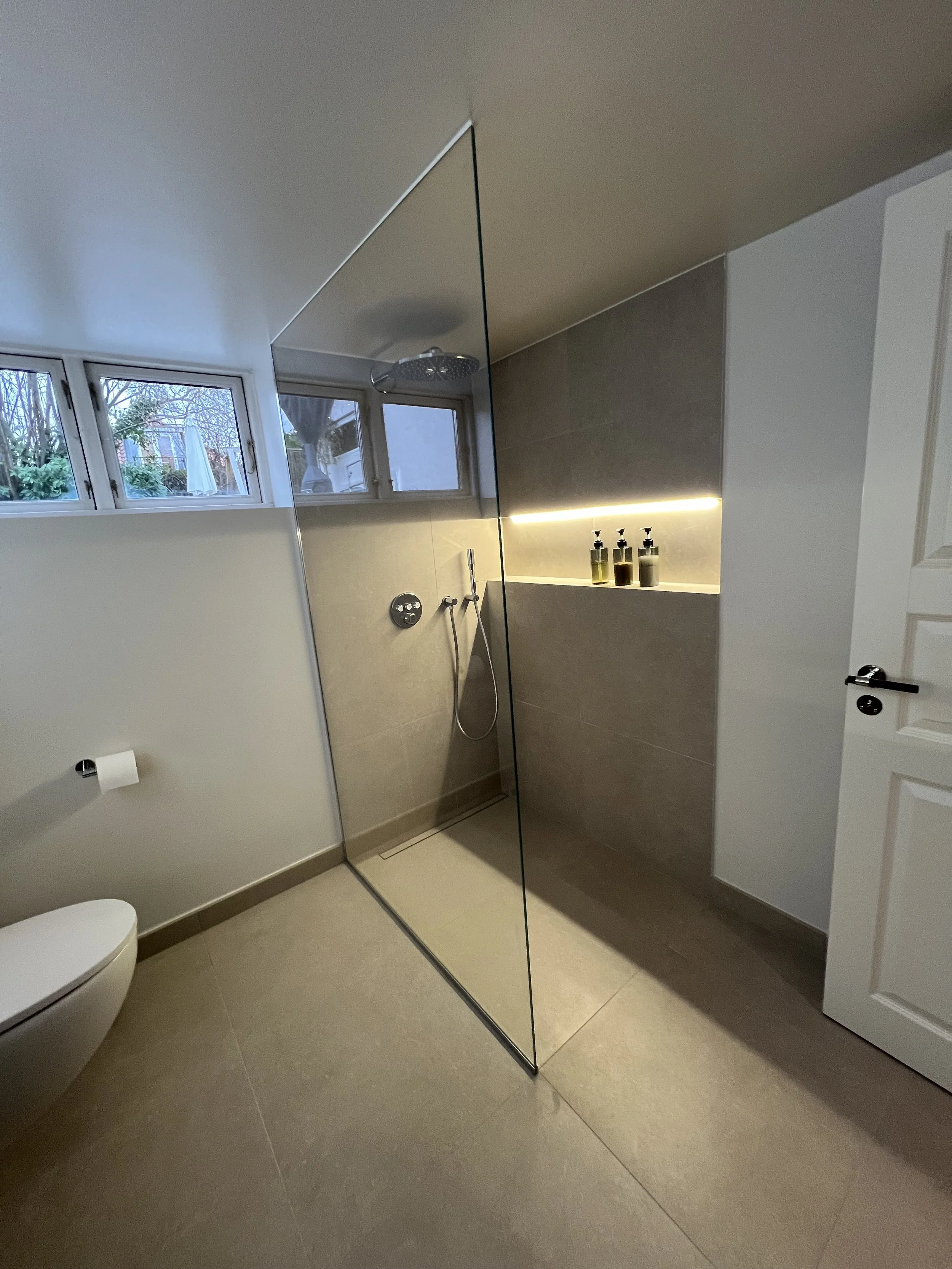 Moderne badeværelse med bruseniche med glasvæg, beige gulv- og vægfliser, tre små vinduer, toilet, oplyst nichesamling med flydende hylder og sæbe- eller shampoo-flasker.