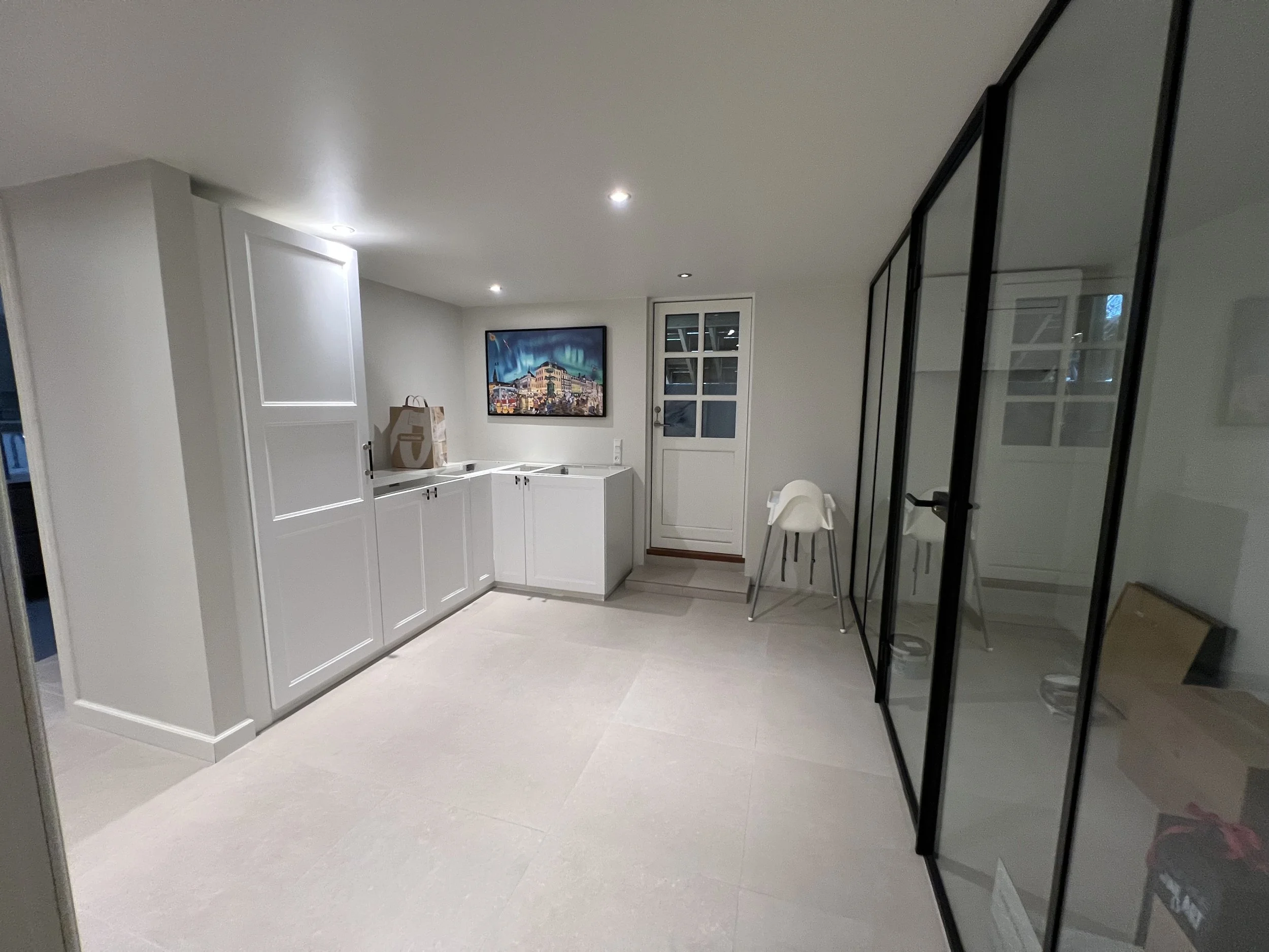 Et tomt rum med hvide skabe og et glas skab med sorte rammer. Der er en glasdør, et maleri på væggen, og en højstol. Rummet har etableret belysning i loftet.