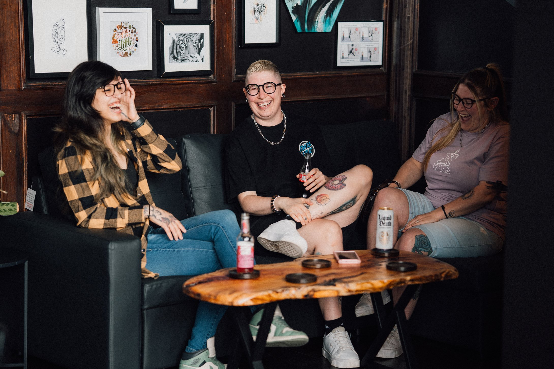 Three women sitting on a black leather couch in a room with framed artwork on dark wood walls having a consultation, laughing and enjoying each other's company, with drinks on a wooden coffee table in front of them.