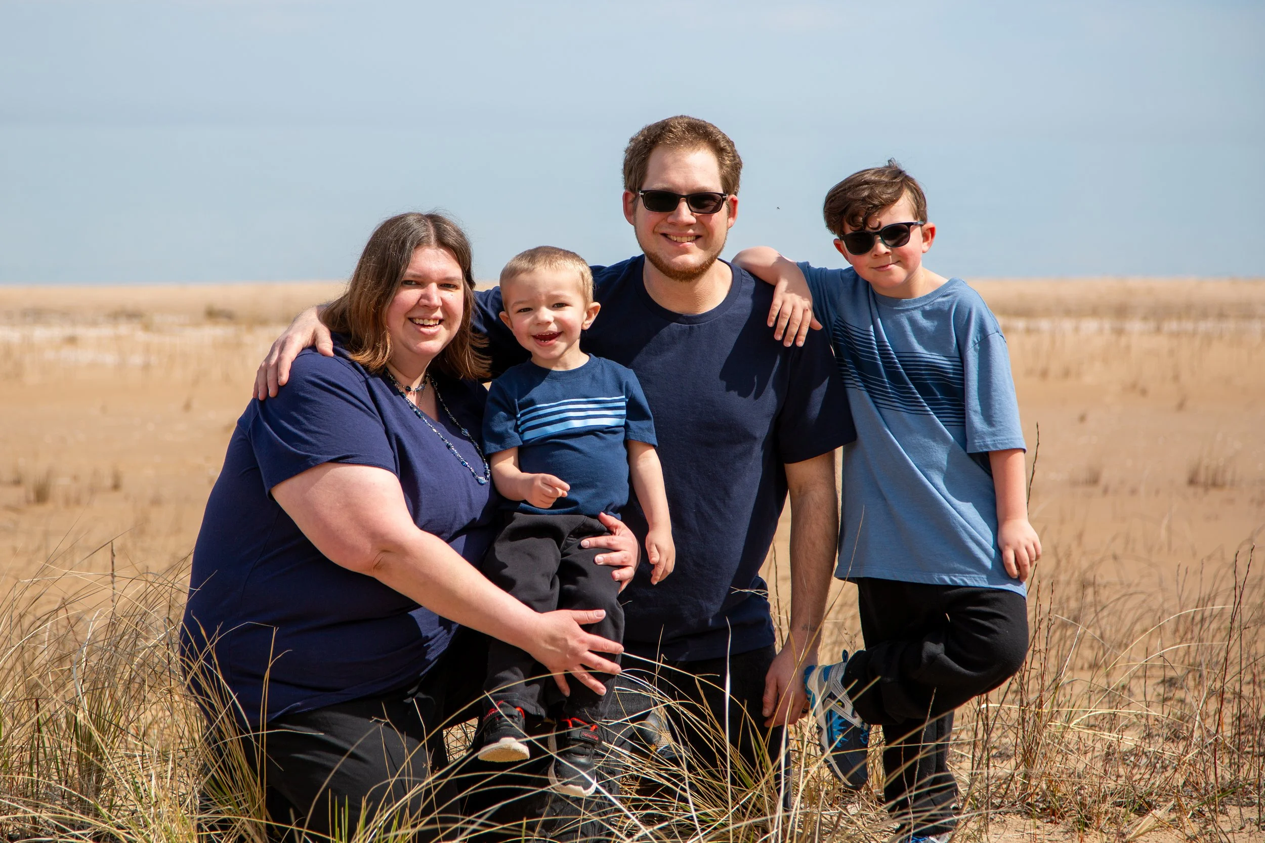 A family of five posing outdoors on a grassy field with a clear sky in the background. The family members are smiling and wearing sunglasses, with the parents kneeling and the children standing, all dressed in casual blue clothing.