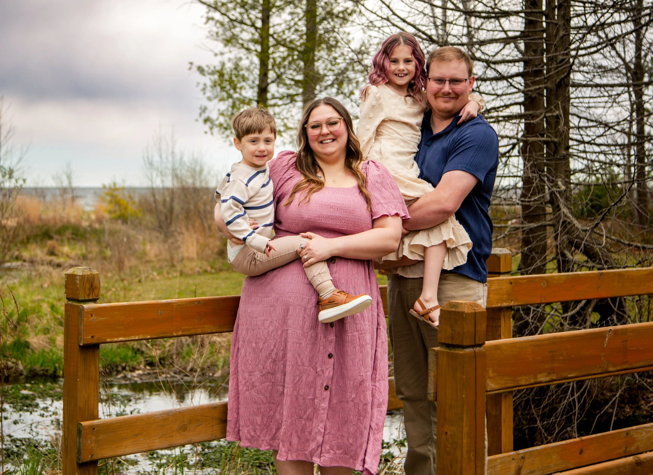 A family of four standing outdoors on a wooden bridge, with trees and cloudy sky in the background. The mother is holding a young boy, and the father is holding a young girl. All are smiling.