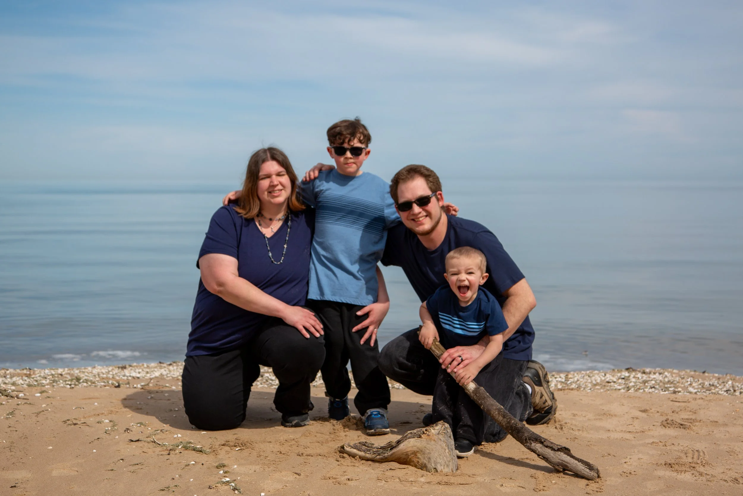 A family of five at the beach, smiling and posing for a photo. They are kneeling and standing on sandy shore near calm water and a partly cloudy sky.