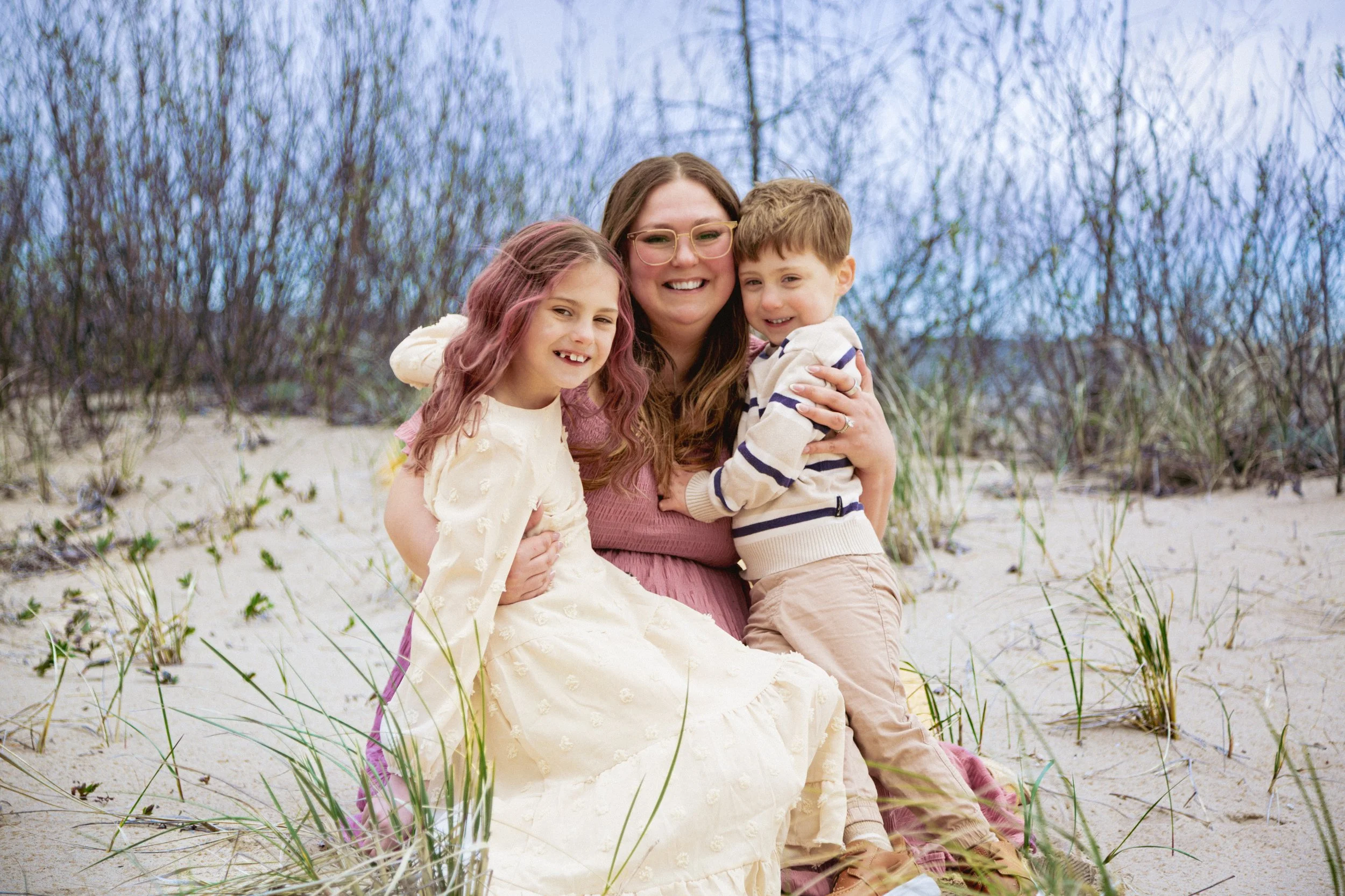A woman with glasses sitting on the beach hugging two children, a girl and a boy, with a background of trees and cloudy sky.