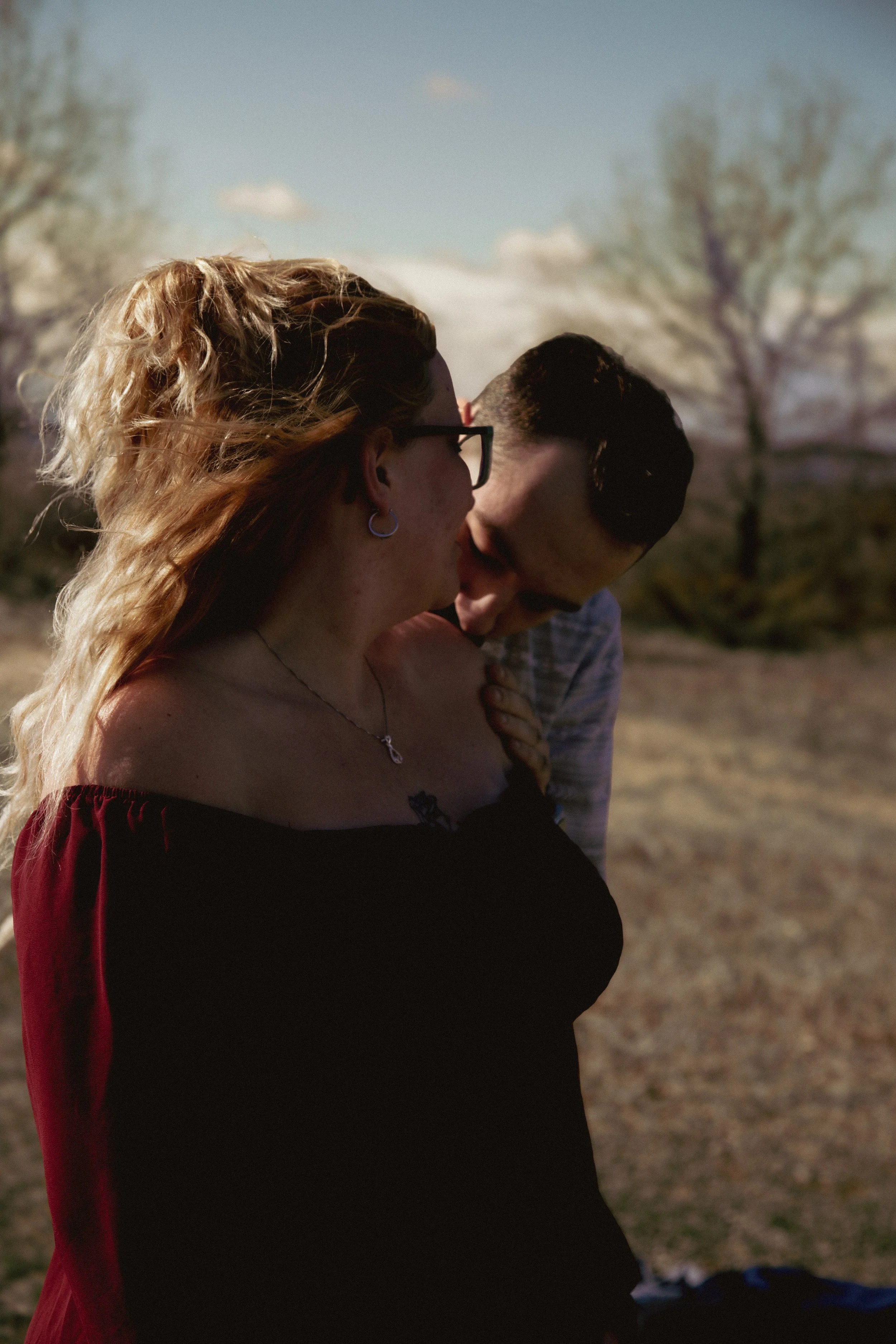 A woman with blonde hair, glasses, a nose ring, and earrings, wearing a off-the-shoulder red top, and a man with short dark hair and facial hair, wearing a striped shirt, sharing an intimate moment outdoors on a sunny day with trees and a blue sky in