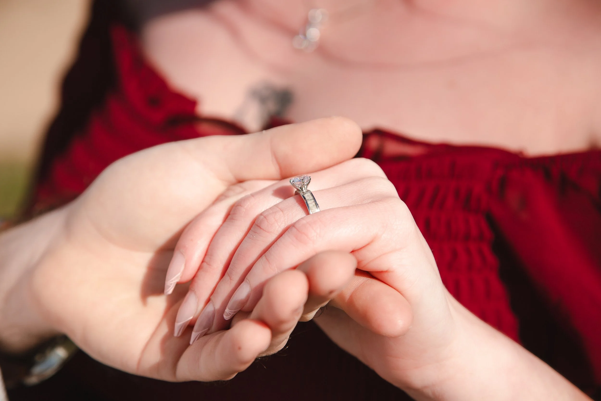 A woman holding a diamond engagement ring in her hand, wearing a red sweater and a necklace.