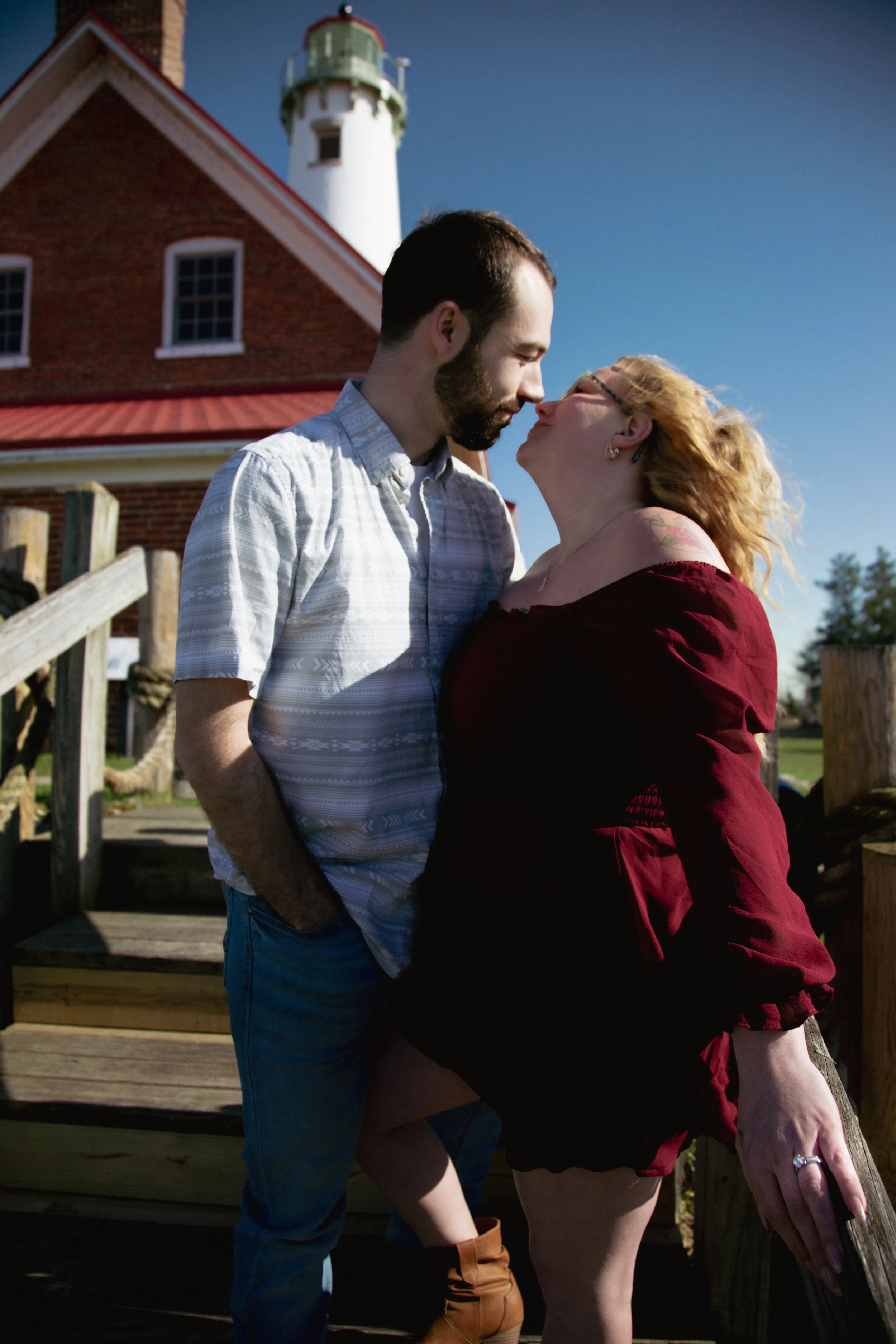 A man and woman standing close together outdoors in front of a red brick lighthouse, leaning their foreheads together. The man wears a light-colored short sleeve shirt and jeans, while the woman has blonde curly hair, glasses, and wears a red off-the