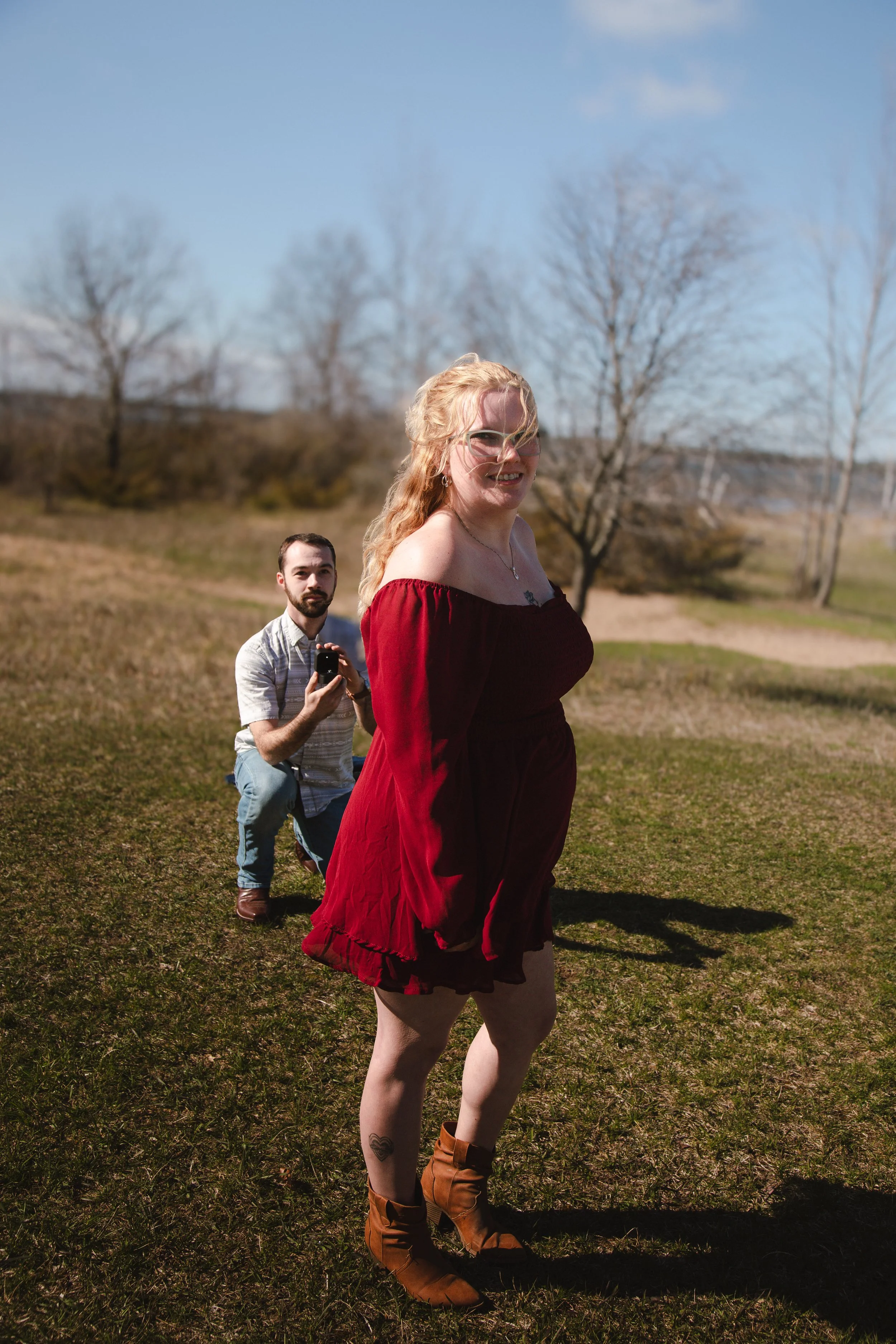 A woman in a red dress and brown boots standing in a grassy field with leafless trees in the background, and a man behind her holding a phone.