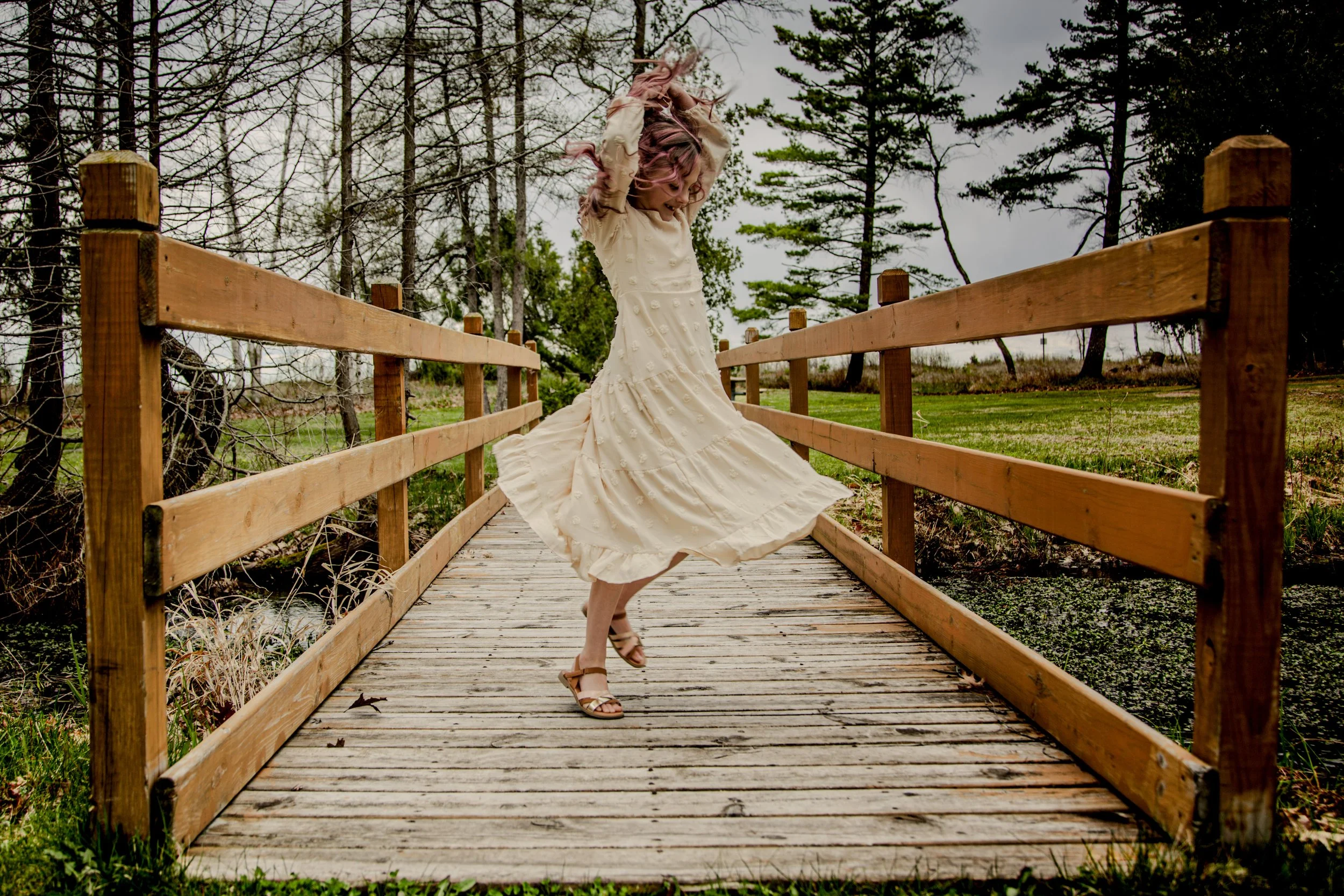 A woman in a white dress with pink hair twirling on a wooden bridge outdoors, surrounded by trees and grass.