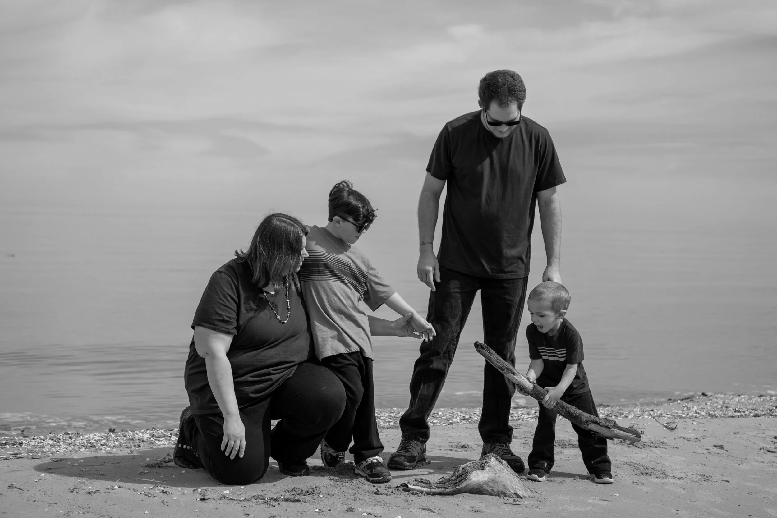 A black and white photo of a family at the beach, with two adults and two children. They are gathered around a piece of driftwood on the sand, looking at something nearby, with an ocean and cloudy sky in the background.