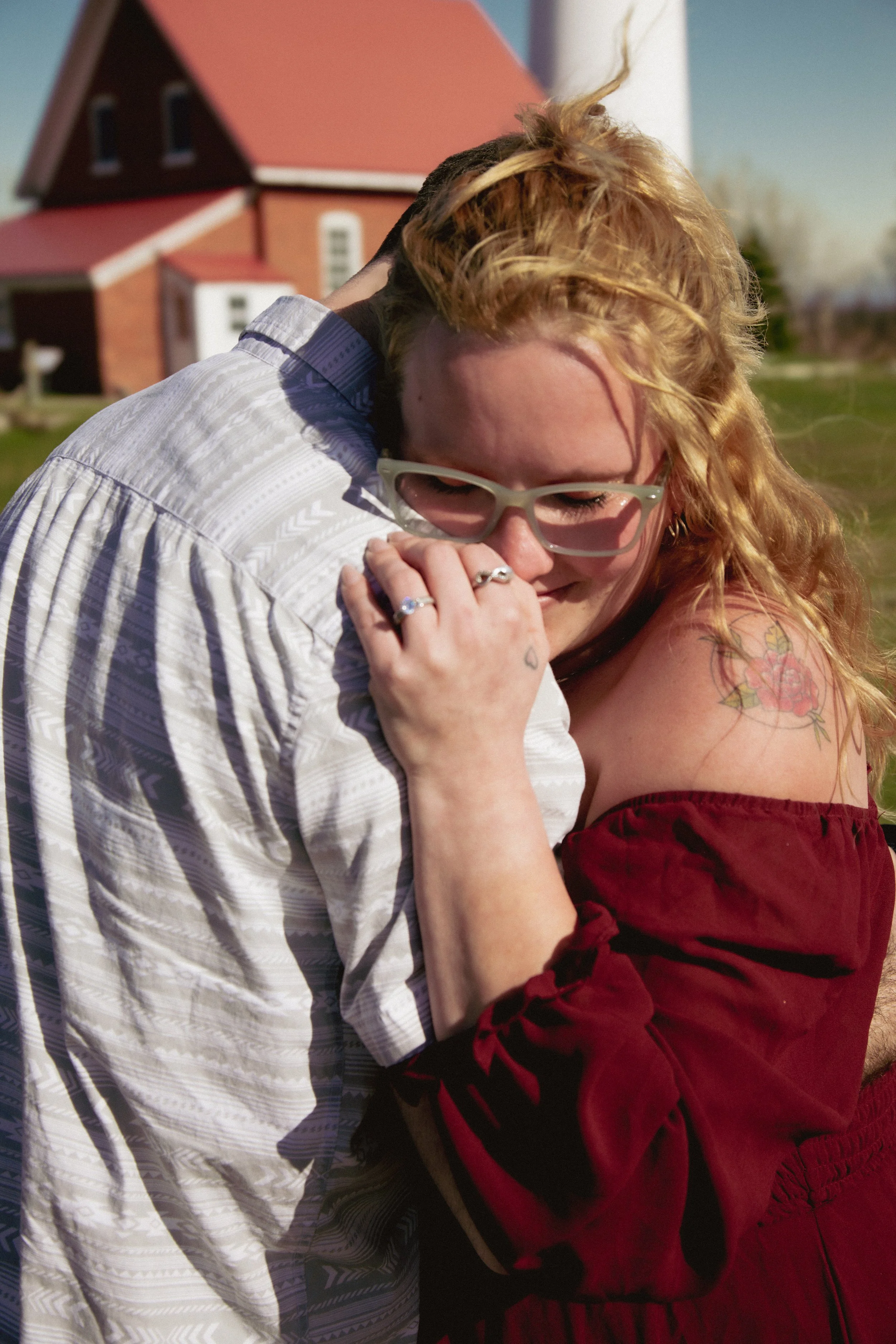 A man and woman hugging outdoors on a sunny day with a red house in the background. The woman has red hair and is wearing glasses and a red dress, while the man has dark hair and is wearing a striped shirt.