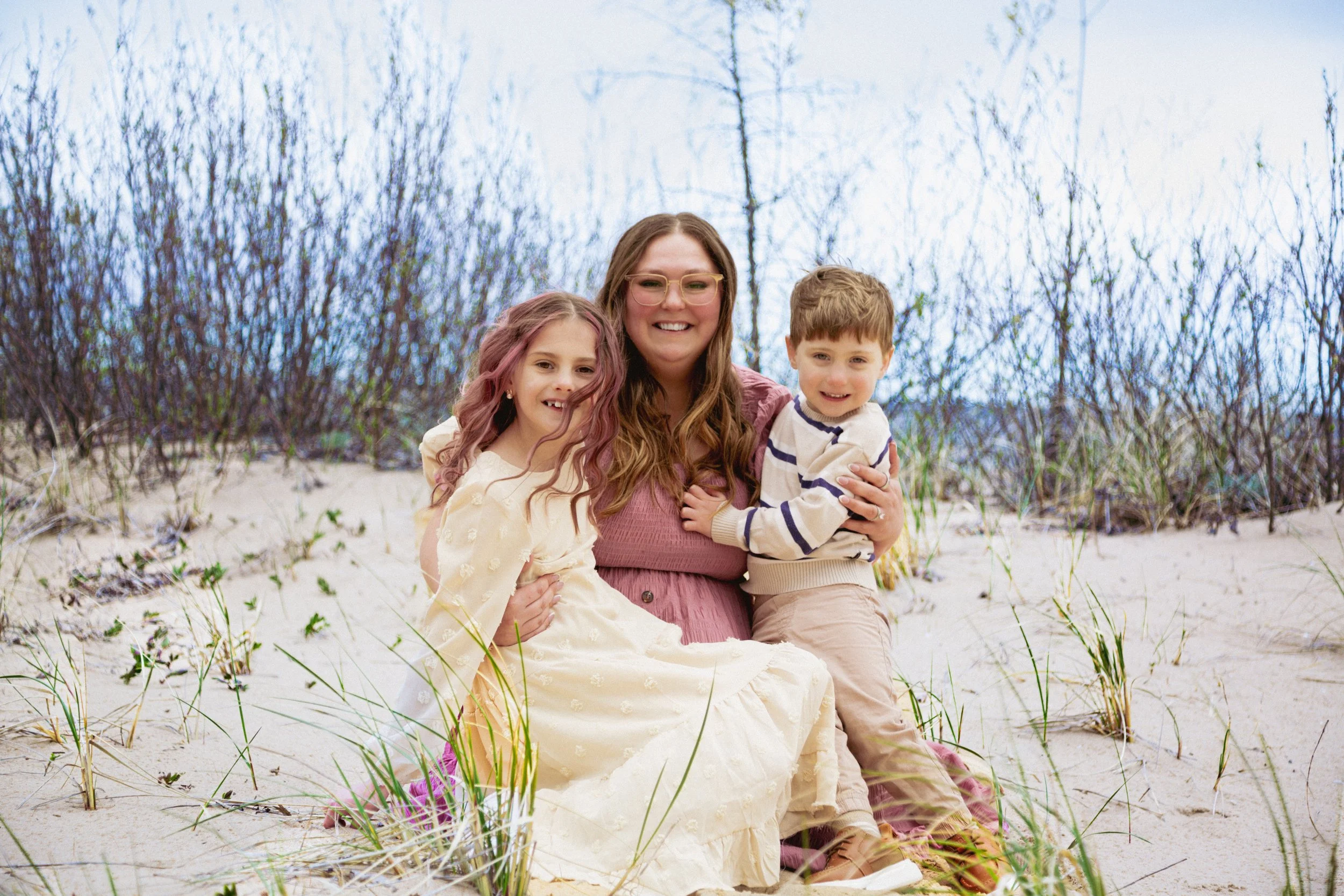 A woman with long hair and glasses smiling while sitting on the sand with two children, a girl with wavy hair and a boy with short hair, surrounded by beach grass and bushes on a sandy beach.