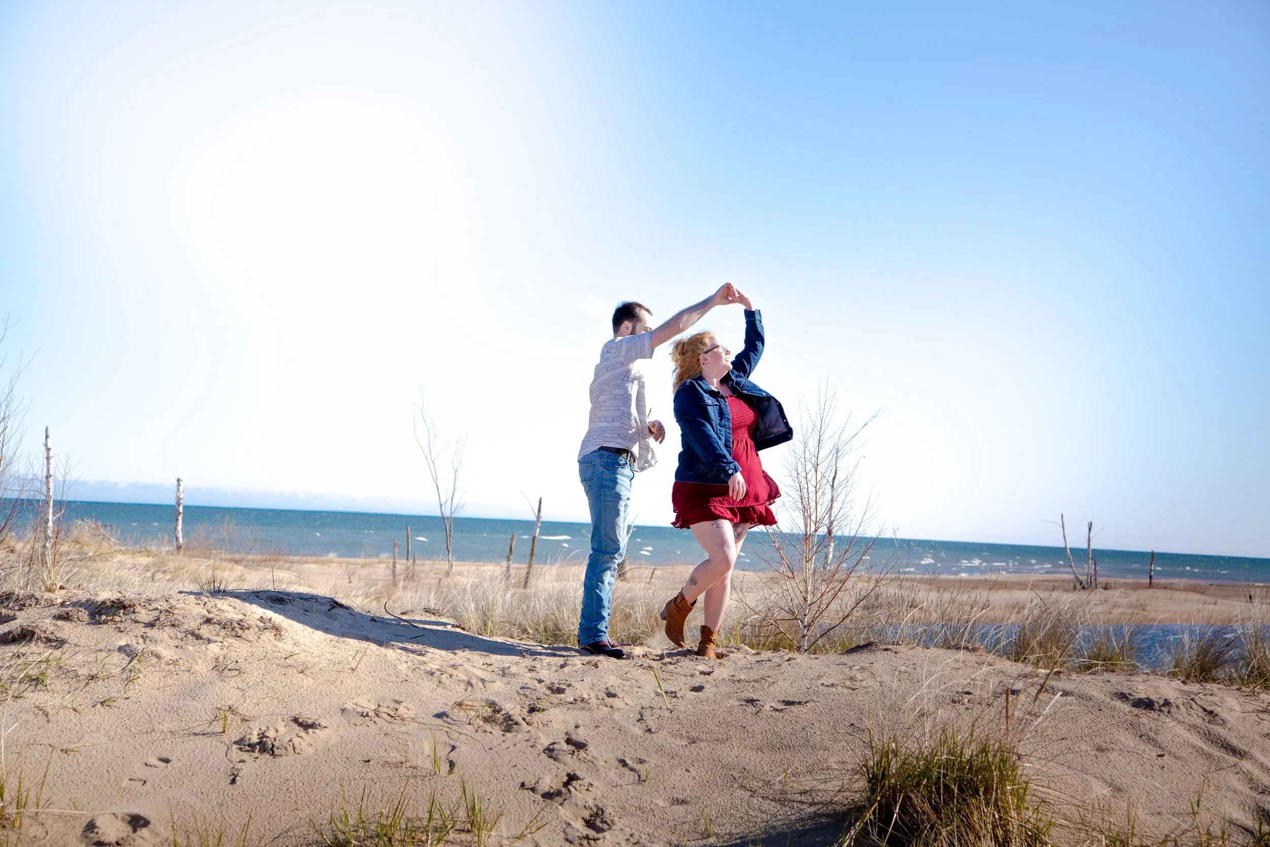 A man and a woman dancing on the beach. The man is holding the woman's hand, and they are spinning together. The woman is wearing a red dress and brown boots, and the man is dressed casually in jeans and a shirt. The beach has sandy dunes with some s