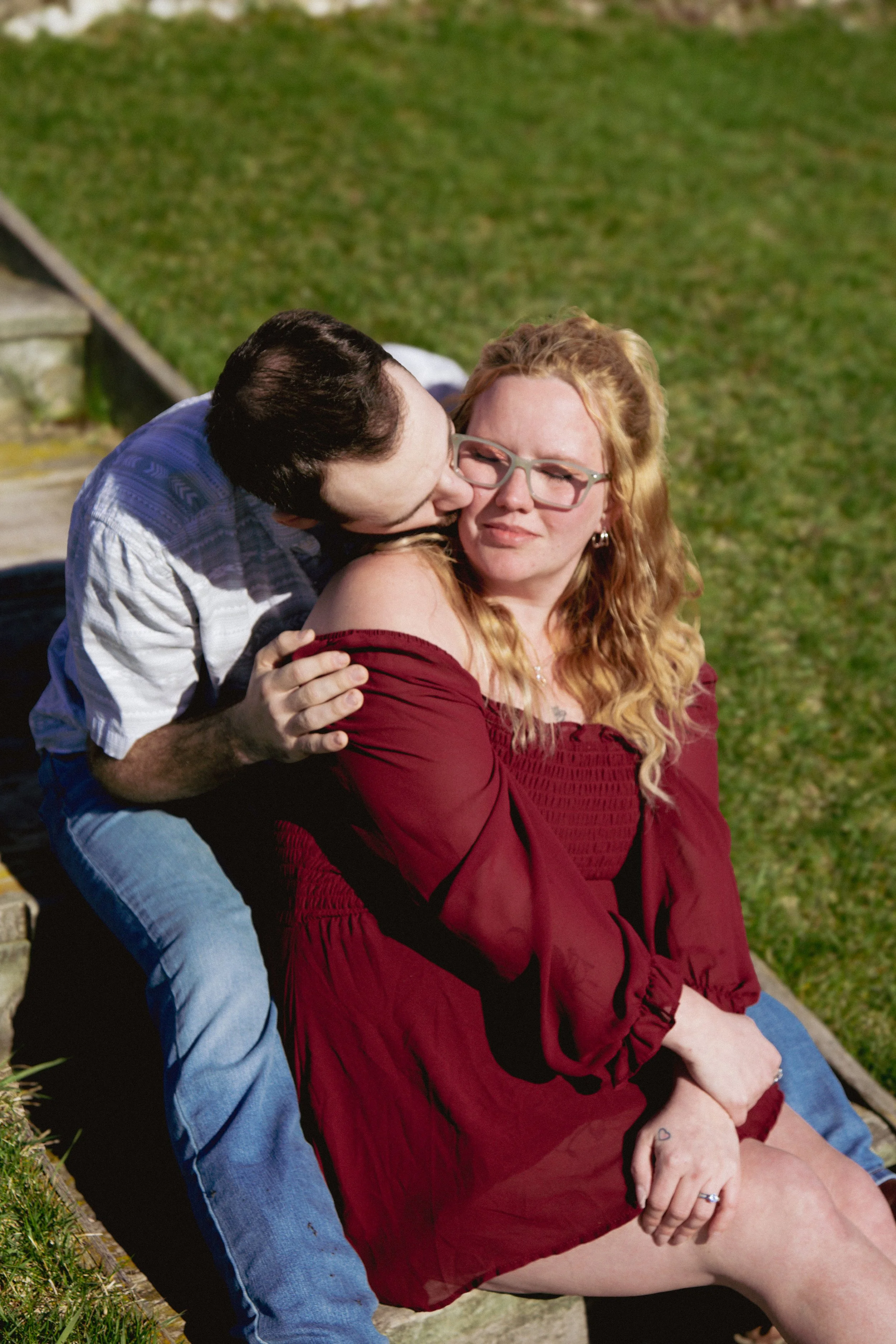 A man giving a woman a kiss on the cheek outdoors, sitting on a park bench. The woman is sitting with her hands on her lap, wearing glasses and a red off-shoulder dress, the man is leaning over from behind, wearing a blue shirt and jeans. The backgro