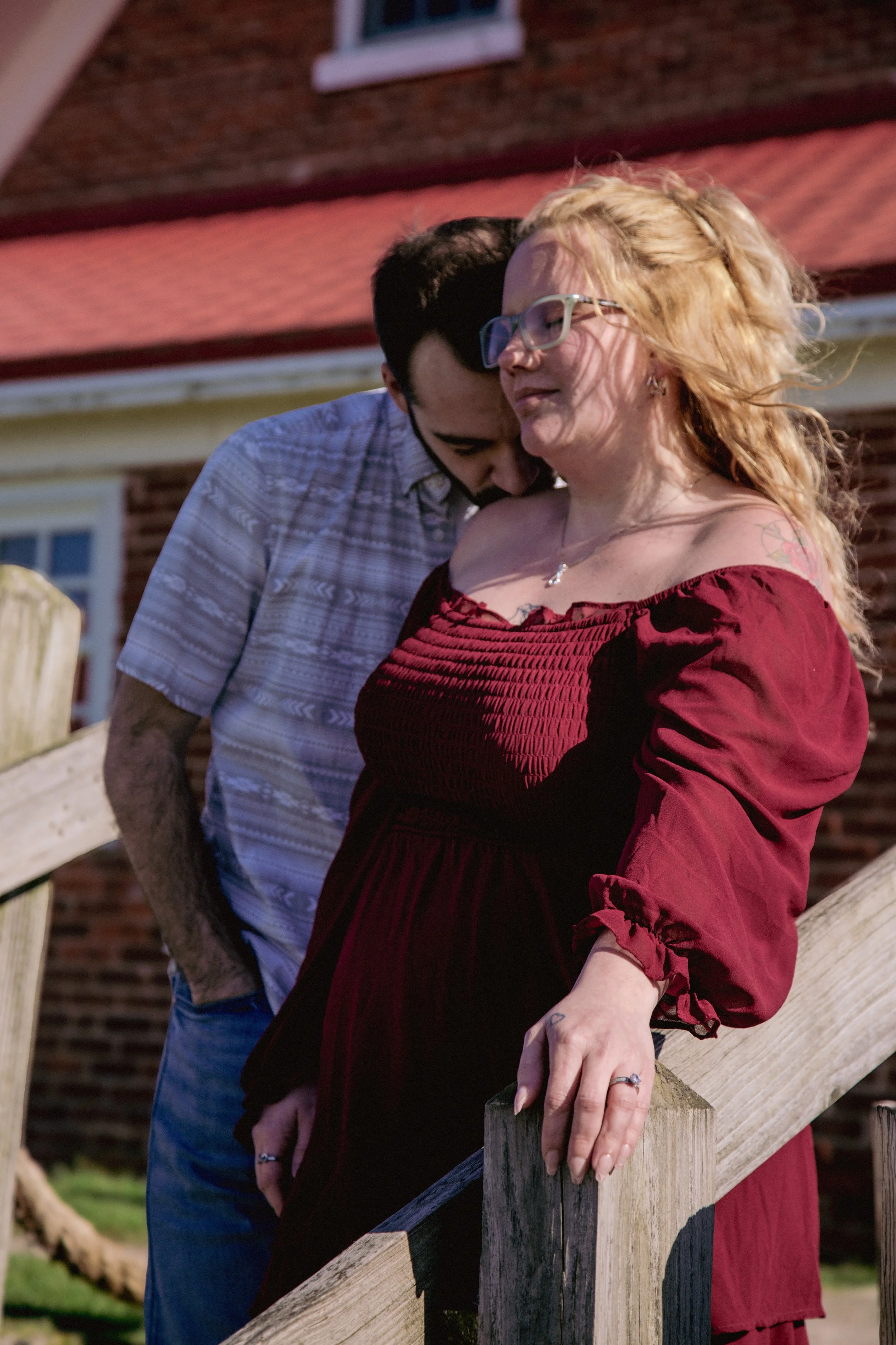 A man and woman share an intimate moment on a wooden staircase outside a brick house, with the woman wearing a red dress and glasses, and the man leaning in close.