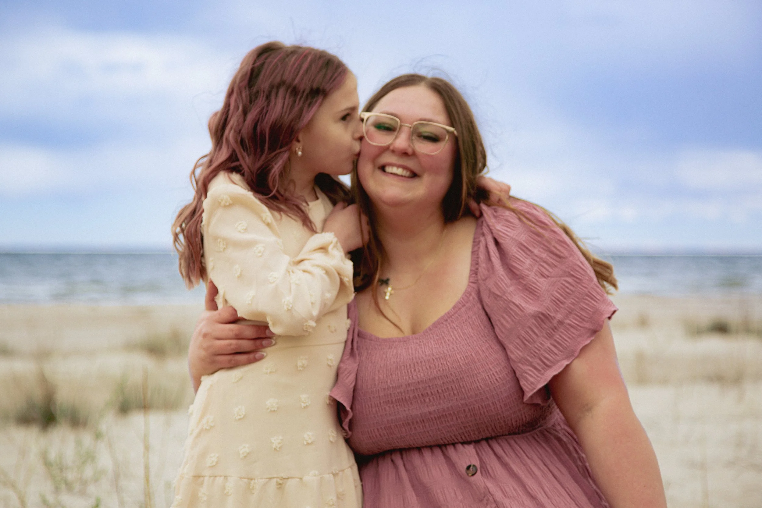 Young woman and girl at the beach, girl kissing woman on cheek, sea and cloudy sky in background.