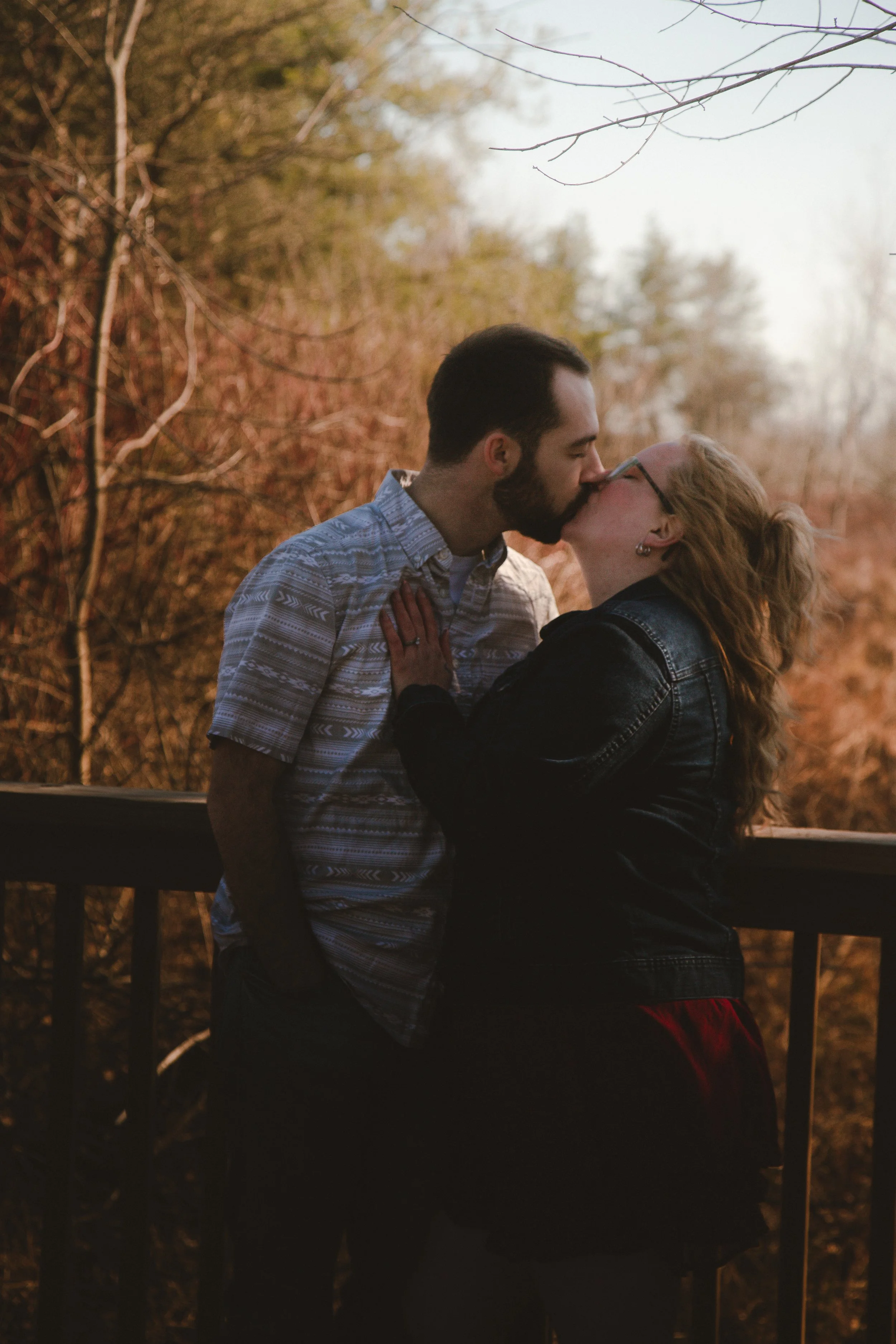 A man and woman sharing a kiss outdoors during sunset, with trees and a wooden fence in the background.
