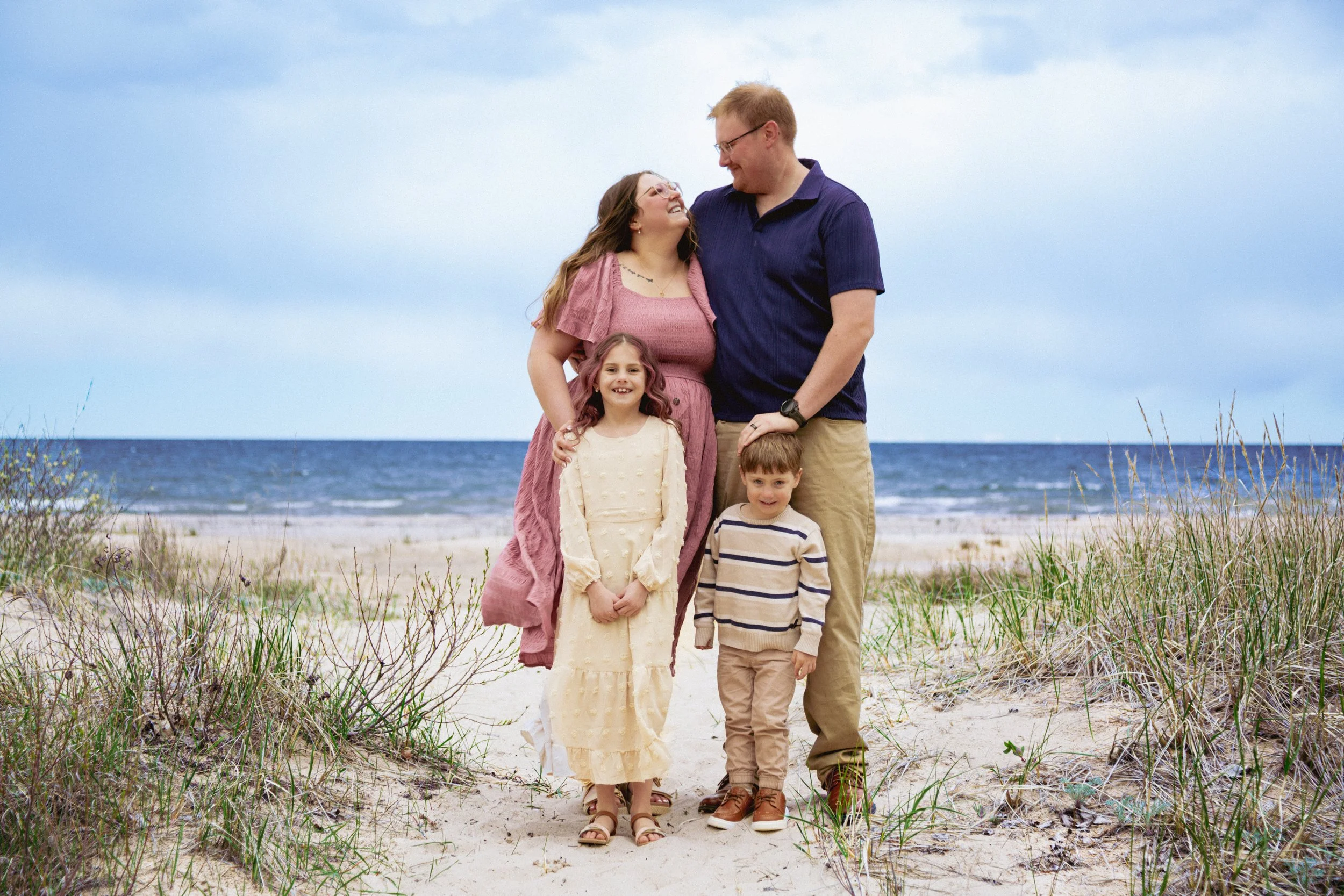 Family of five enjoying a day at the beach, standing on sand among grass with the ocean and cloudy sky in the background. The family includes a woman, a man, and three children, all smiling and dressed casually.