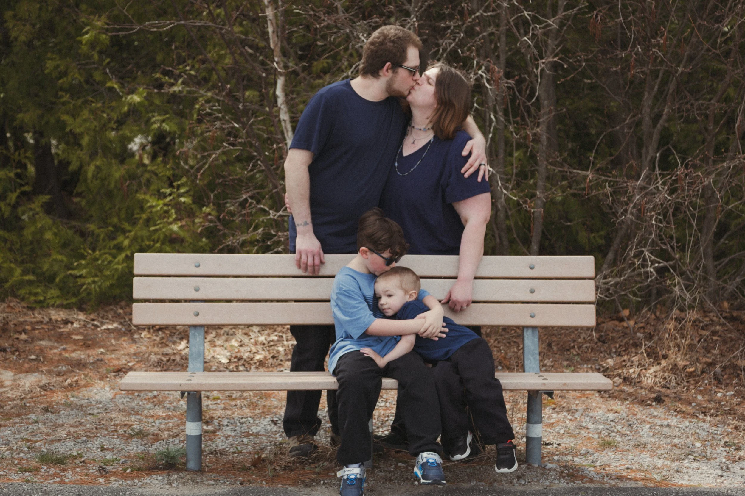 A couple kissing while one of the children is hugging the other on a park bench in front of a background of trees.