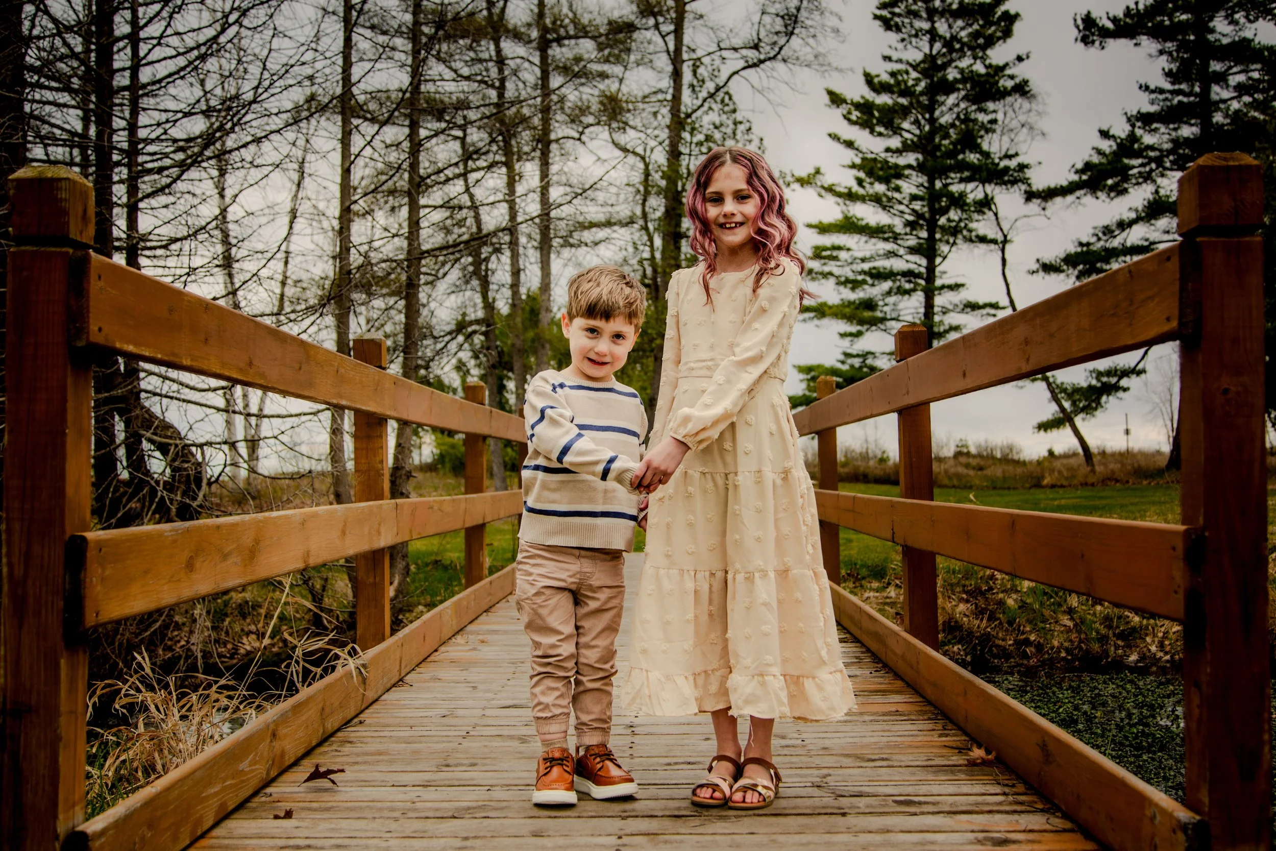A young girl and boy holding hands on a wooden bridge in a wooded area during overcast weather.