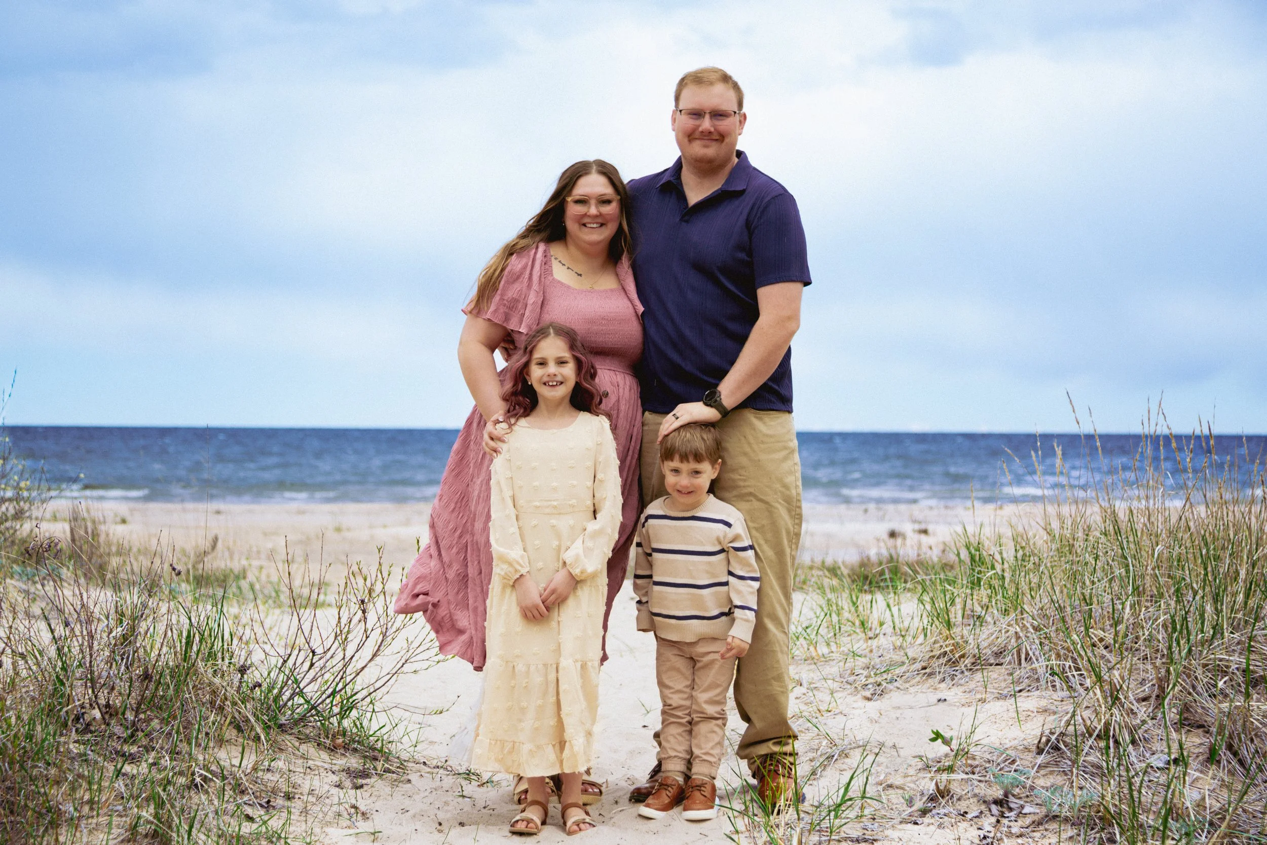 A family of five standing on a sandy beach with grassy dunes in the foreground and the ocean in the background, under a cloudy sky.