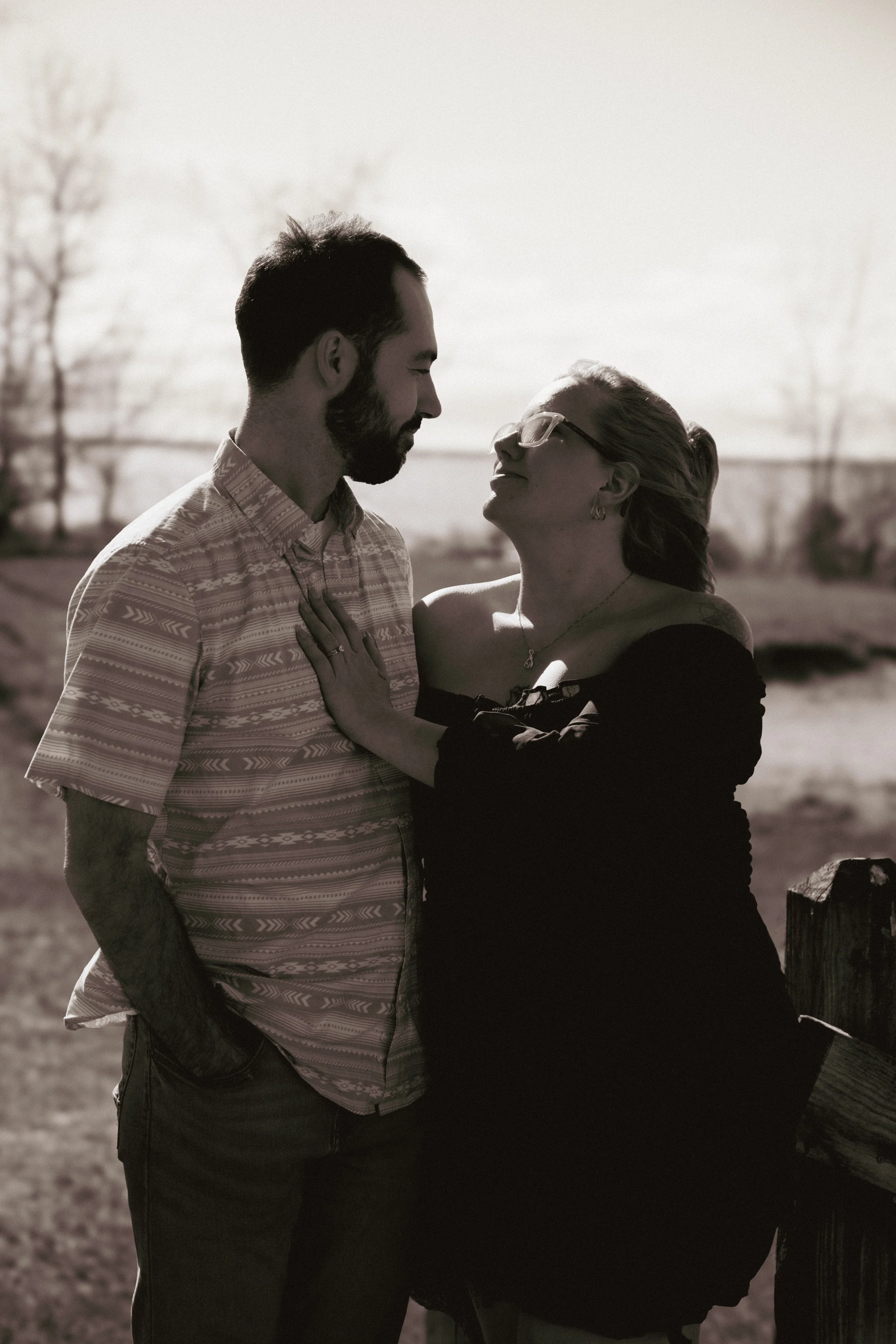 A black and white photo of a man and woman facing each other outdoors, with the man holding the woman's shoulder, both with closed eyes, near a wooden fence with trees in the background.