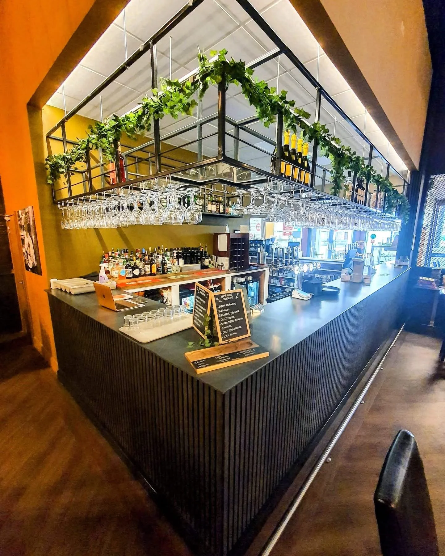 Indoor bar area with hanging glassware, bottles of alcohol on shelves, a menu board, and decorative greenery overhead.