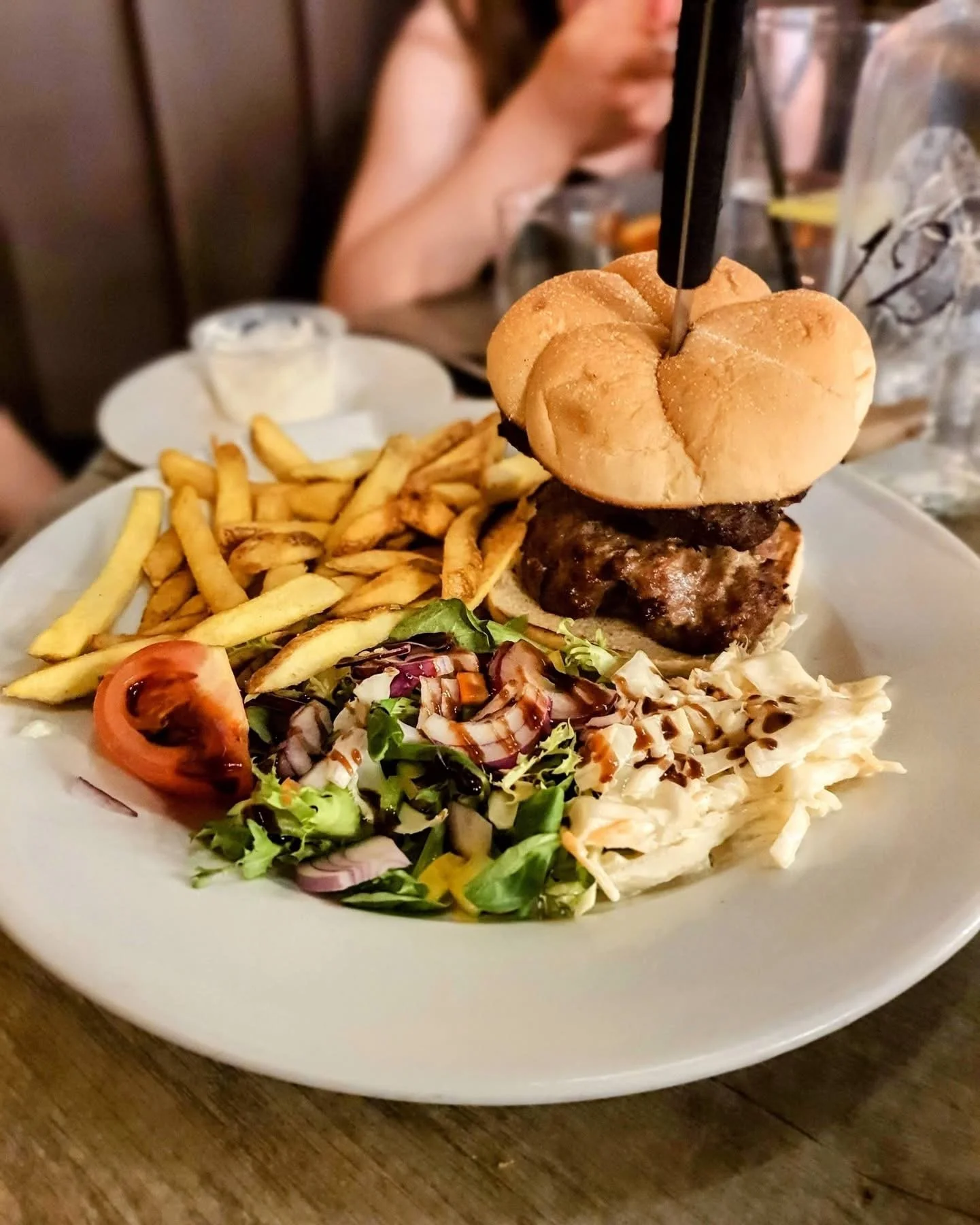 Plate with French fries, a side salad, and a burger with a bun and a beef patty, view of other people eating in the background.