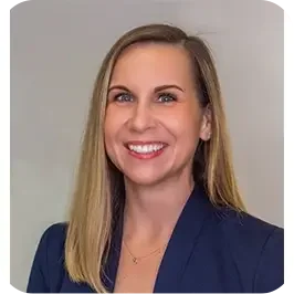 A woman with long blonde hair wearing a dark blazer, smiling against a light-colored background.
