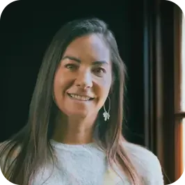 A smiling woman with long brown hair wearing a white top, standing near a window with dark curtains.