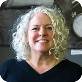 A woman with curly gray hair smiling in a scientific or industrial setting.