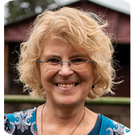 A smiling middle-aged woman with curly blonde hair, glasses, and a blue patterned shirt standing outdoors