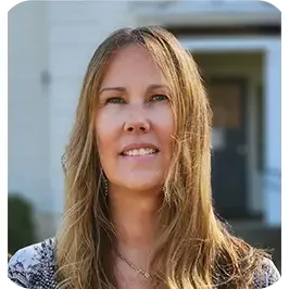 A woman with long light brown hair standing outside in front of a house.