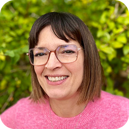 A woman with shoulder-length brown hair and glasses, wearing a pink sweater, smiling outdoors with green foliage in the background.