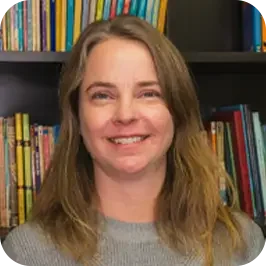 A woman with long, light brown hair, smiling, in front of a bookshelf filled with books.