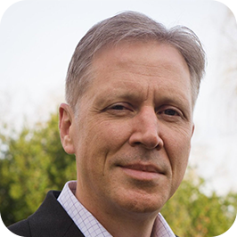 Close-up of a middle-aged man with gray hair outdoors, wearing a black suit jacket and a collared shirt, with trees in the background.