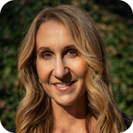 A smiling woman with shoulder-length blonde hair outdoors with green foliage in the background.
