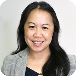 Portrait of a smiling woman with long black hair, wearing a light gray blazer and navy top, standing against a plain light background.