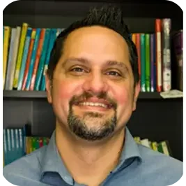 Headshot of a smiling man with short dark hair and a goatee, standing in front of a bookshelf filled with colorful books.
