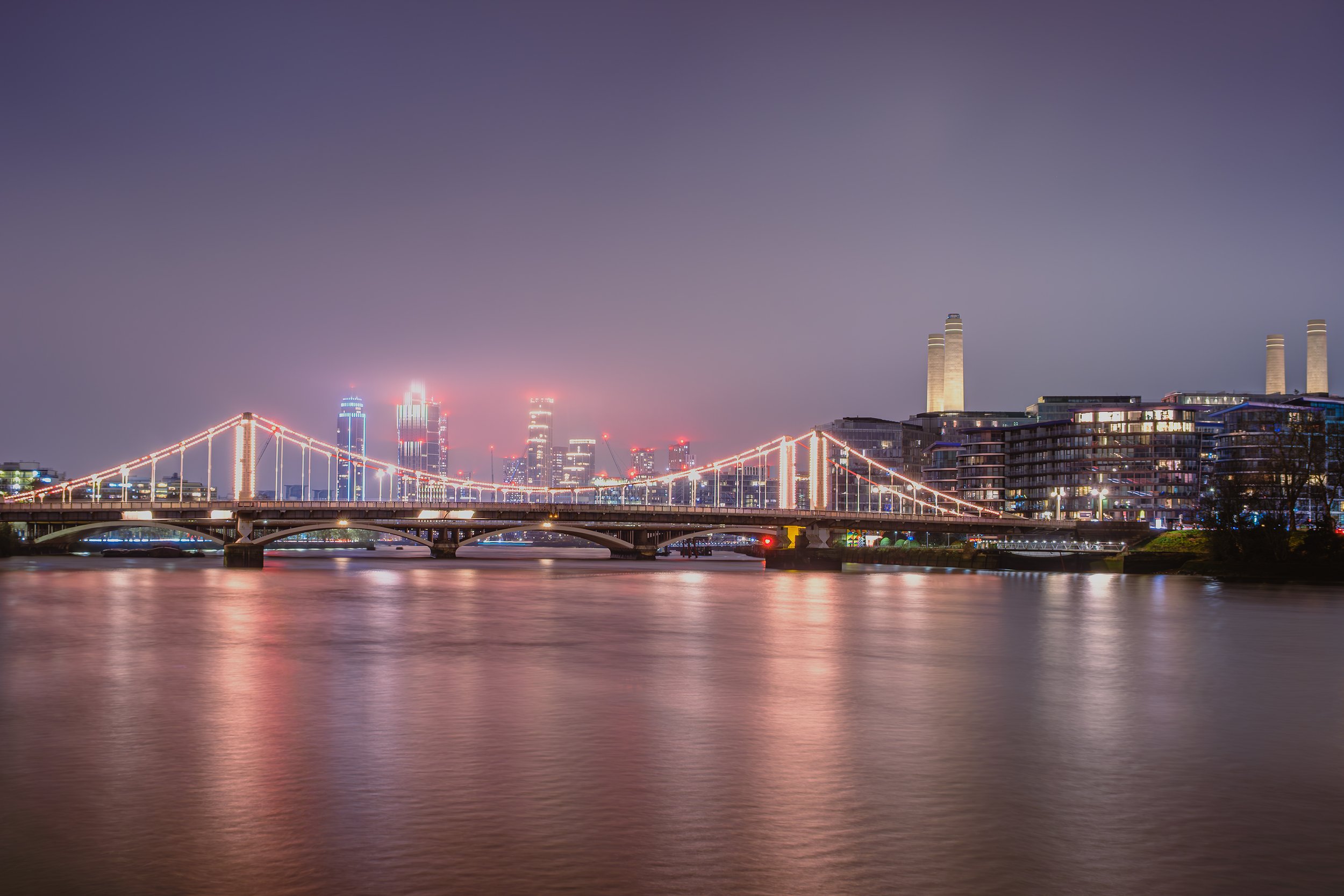 Chelsea Bridge illuminated at blue hour, with the Battersea skyline reflected in the calm waters of the River Thames. The warm lights of the bridge contrast with the soft evening sky, creating a peaceful London cityscape.
Long - exposure photograph c