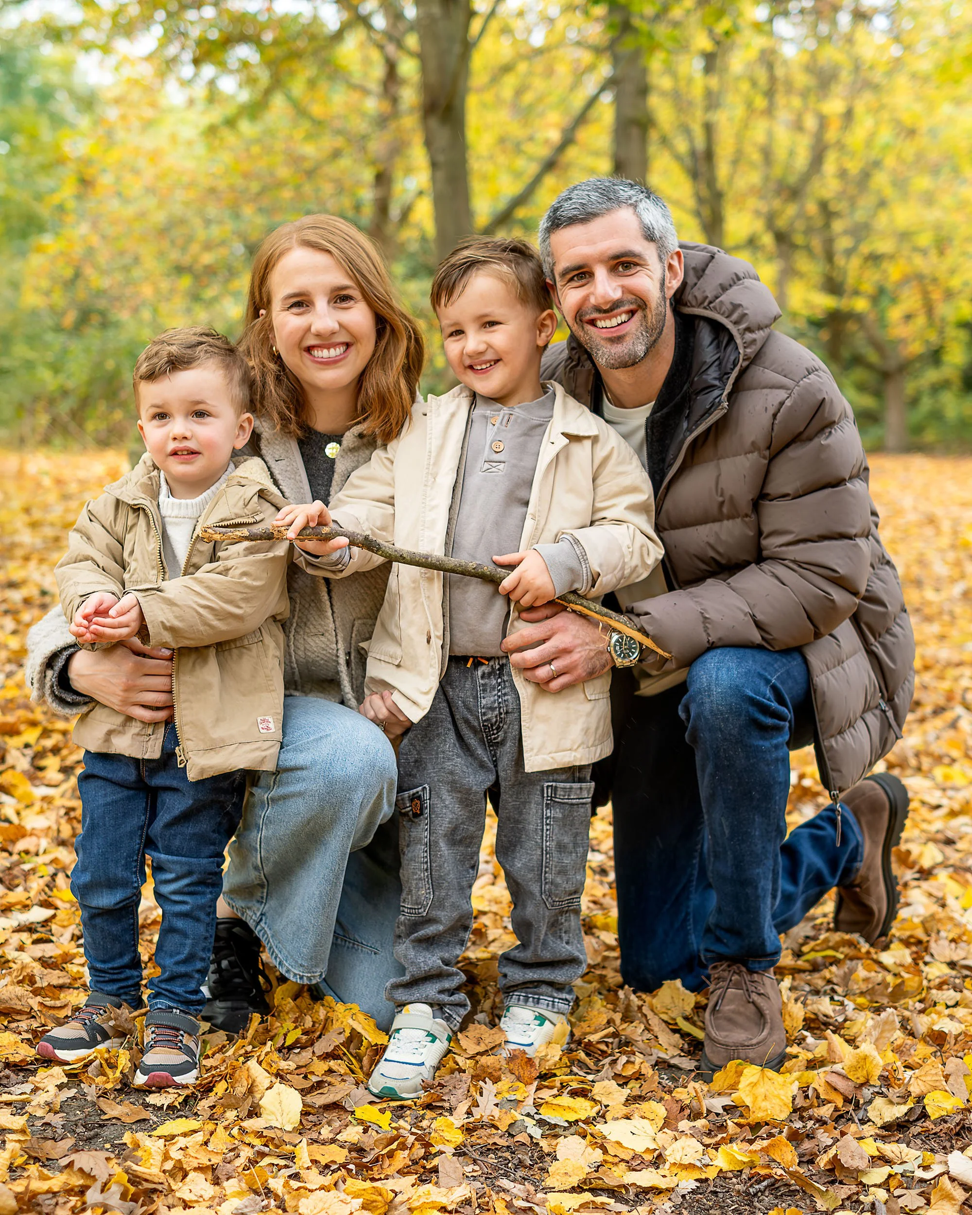Four people, two adults and two children, smiling and holding a stick while outdoors in a park during fall with yellow leaves.