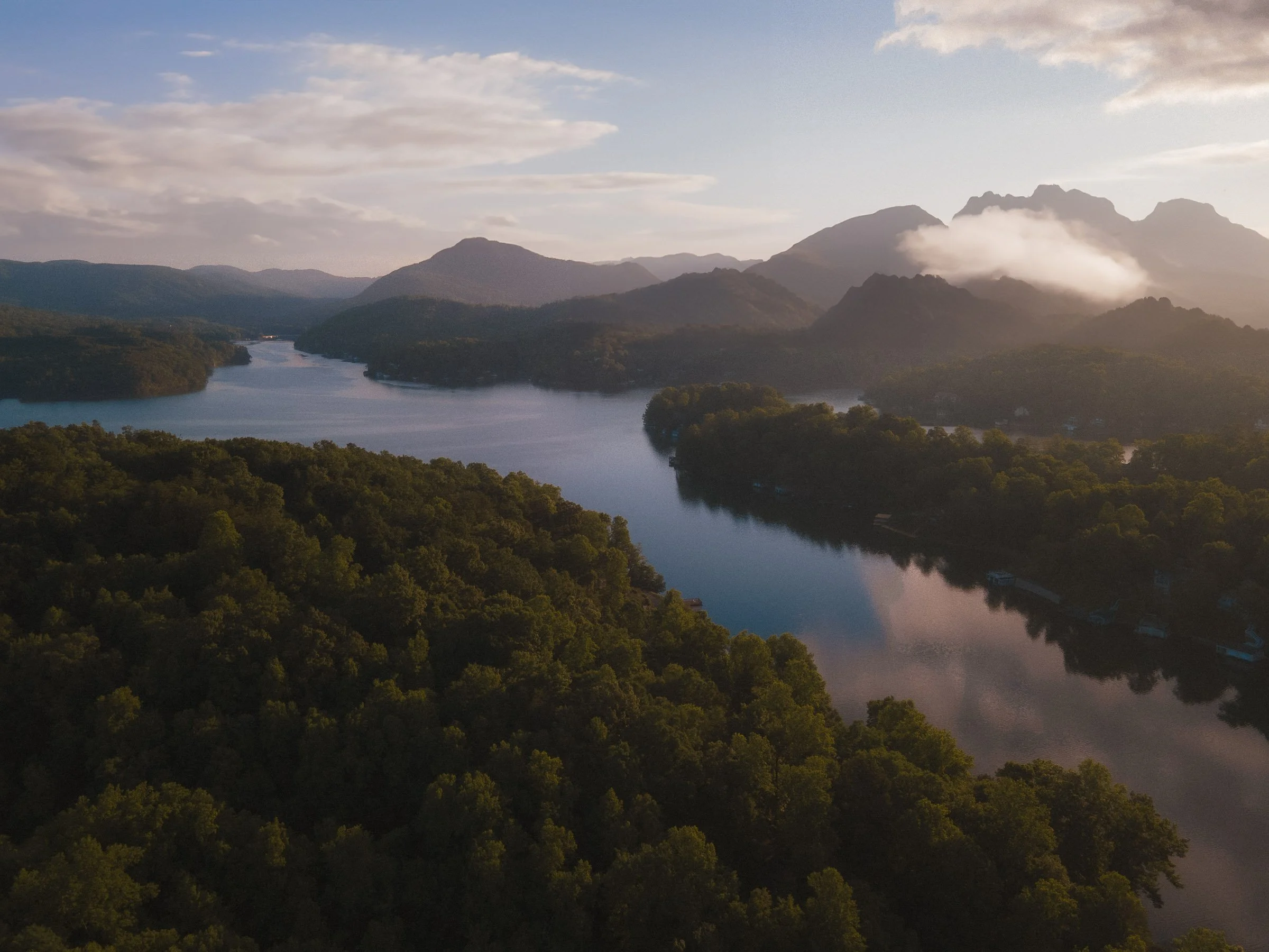 Aerial view of a river winding through a forested landscape with mountains in the background covered in clouds, under a partly cloudy sky.