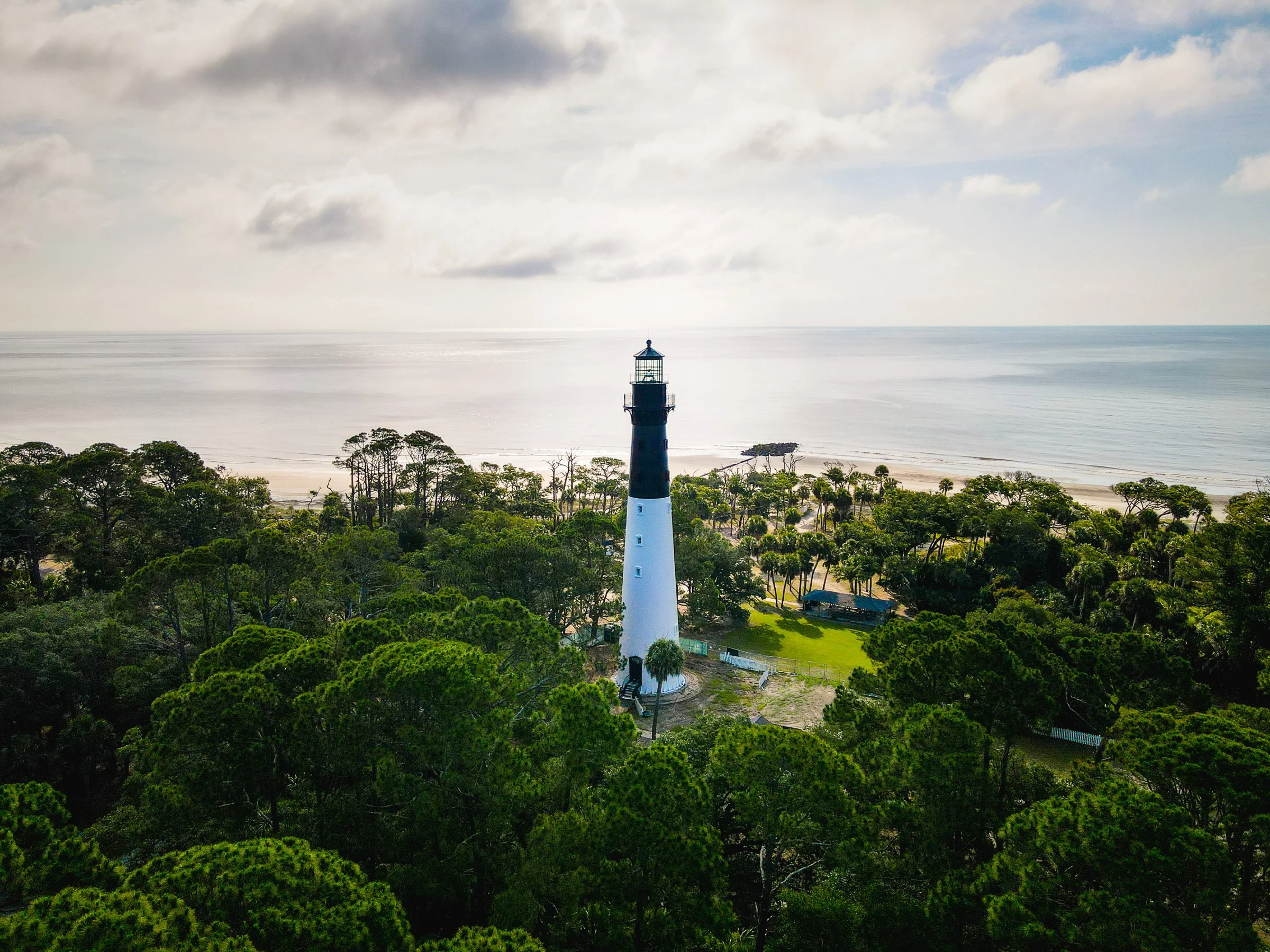 A lighthouse surrounded by lush green trees, with the ocean and cloudy sky in the background.