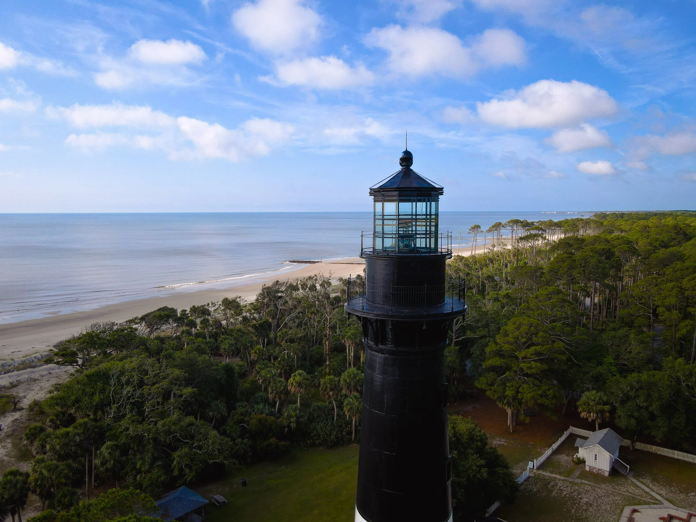 A black lighthouse with glass windows on top of a grassy area near a sandy beach and the ocean, under a partly cloudy sky.