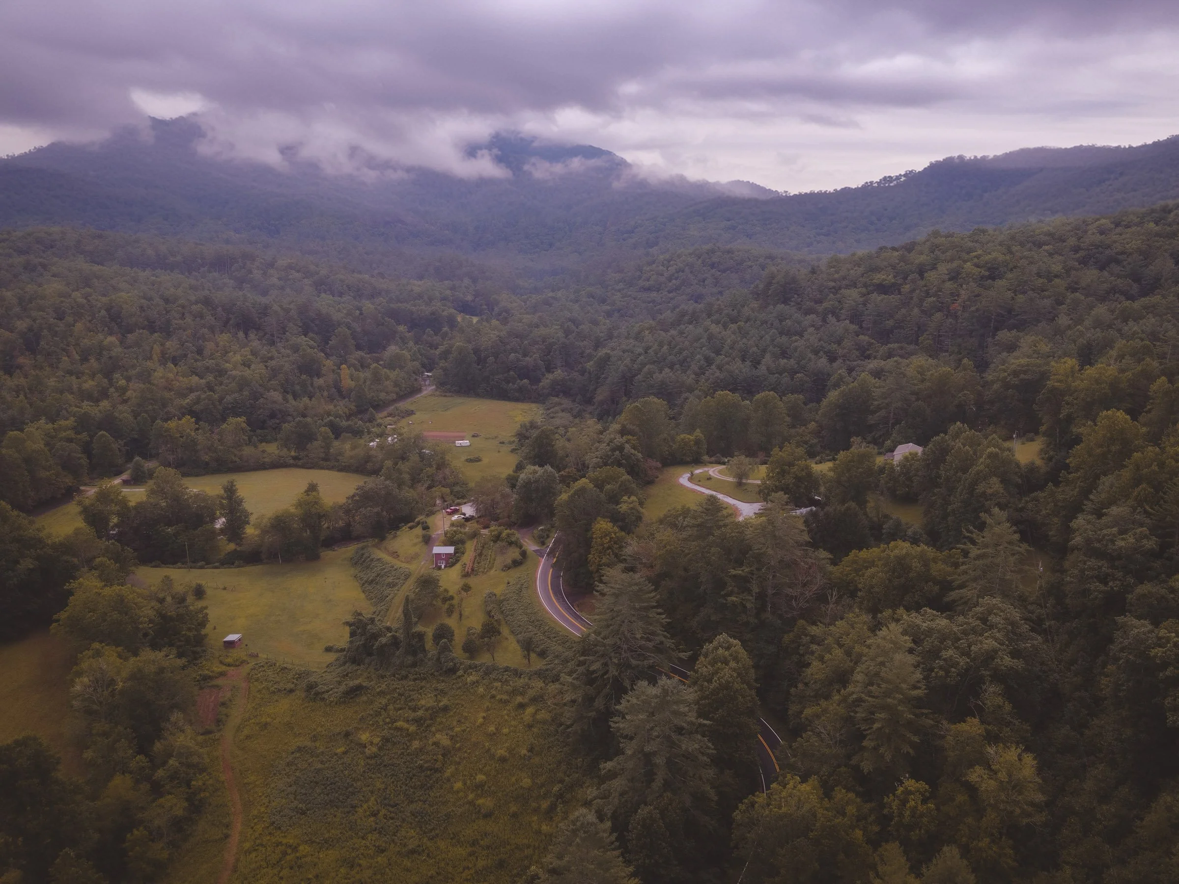 Aerial view of a winding road through a lush green forested mountain landscape with cloudy skies.