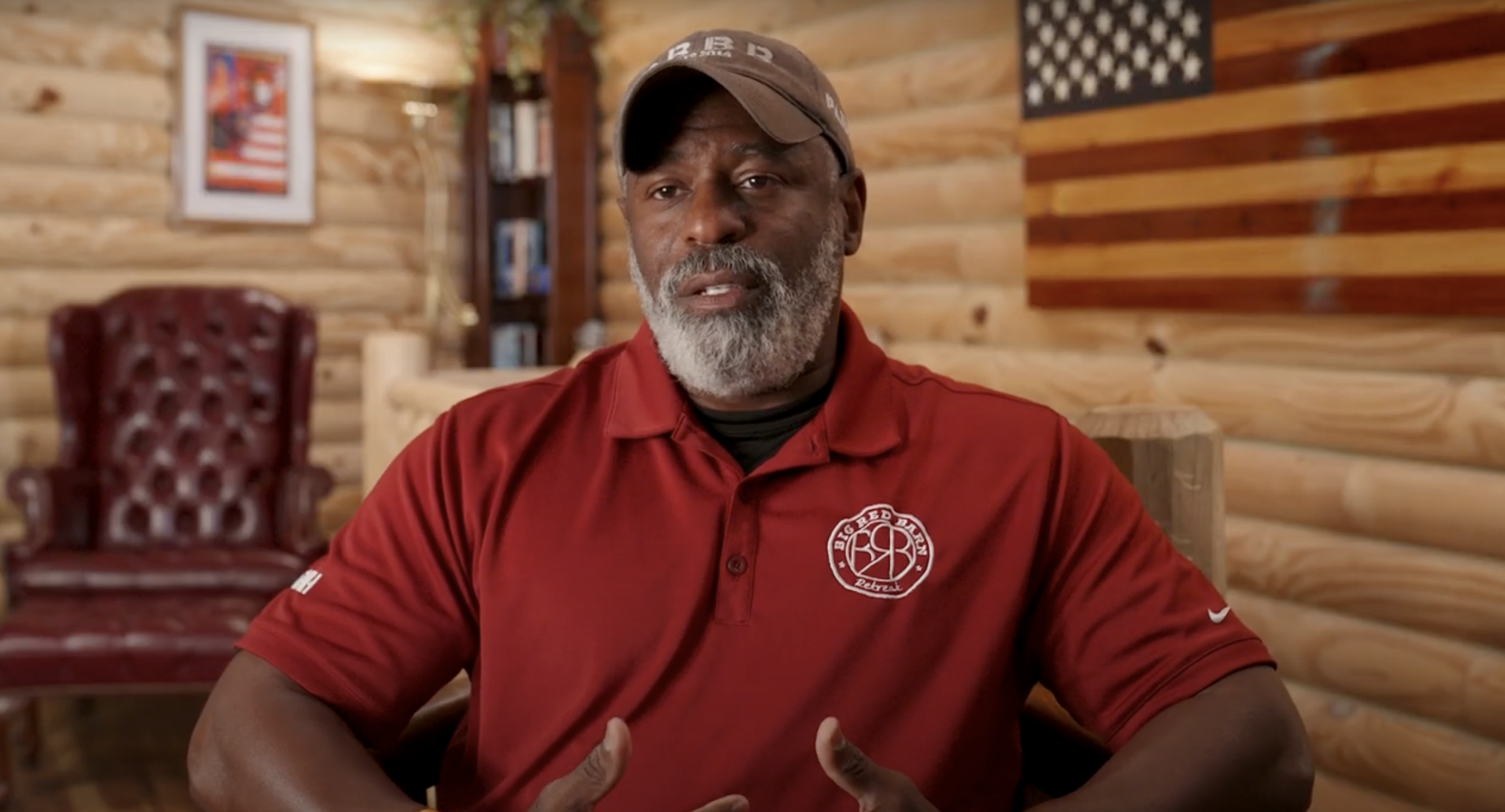 A man with gray hair and beard wearing a red shirt and baseball cap, sitting in a log cabin-style room with wooden walls, a red leather chair, a framed picture on the wall, and an American flag decoration.