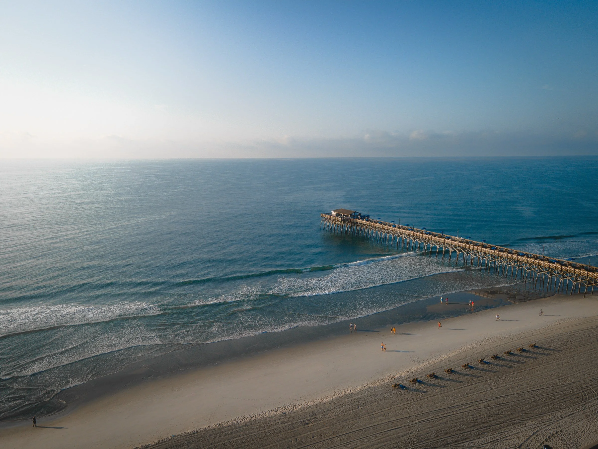 A sandy beach with people walking, biking, and relaxing. A pier extends into the ocean, with a small building at the end, under a blue sky with some clouds.