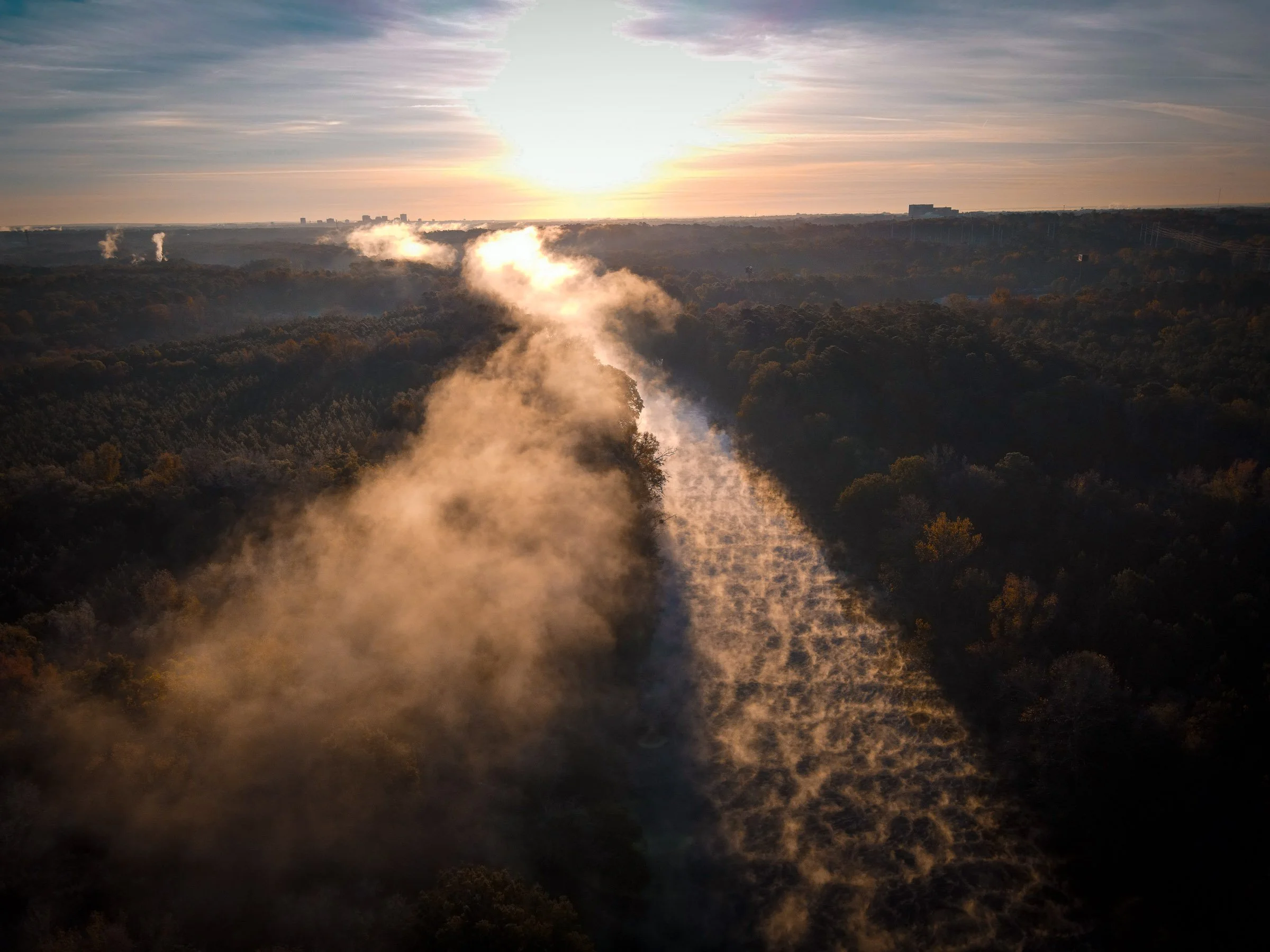 Aerial view of a forest with two rivers in a V-shape, emitting steam or smoke, during sunset with clouds in the sky.