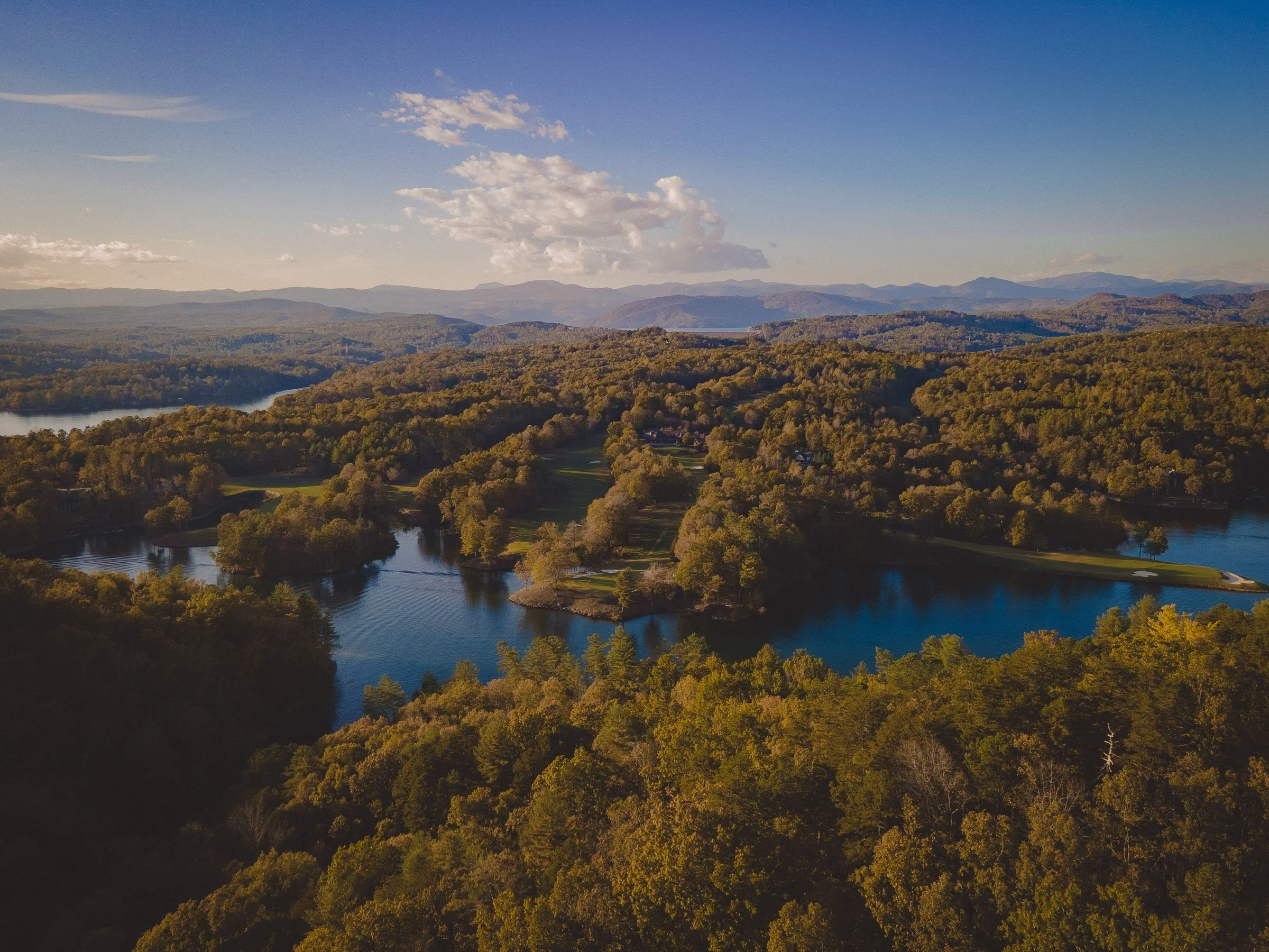 Aerial view of a landscape with a lake, dense forests, hills, and mountains in the distance under a partly cloudy sky.